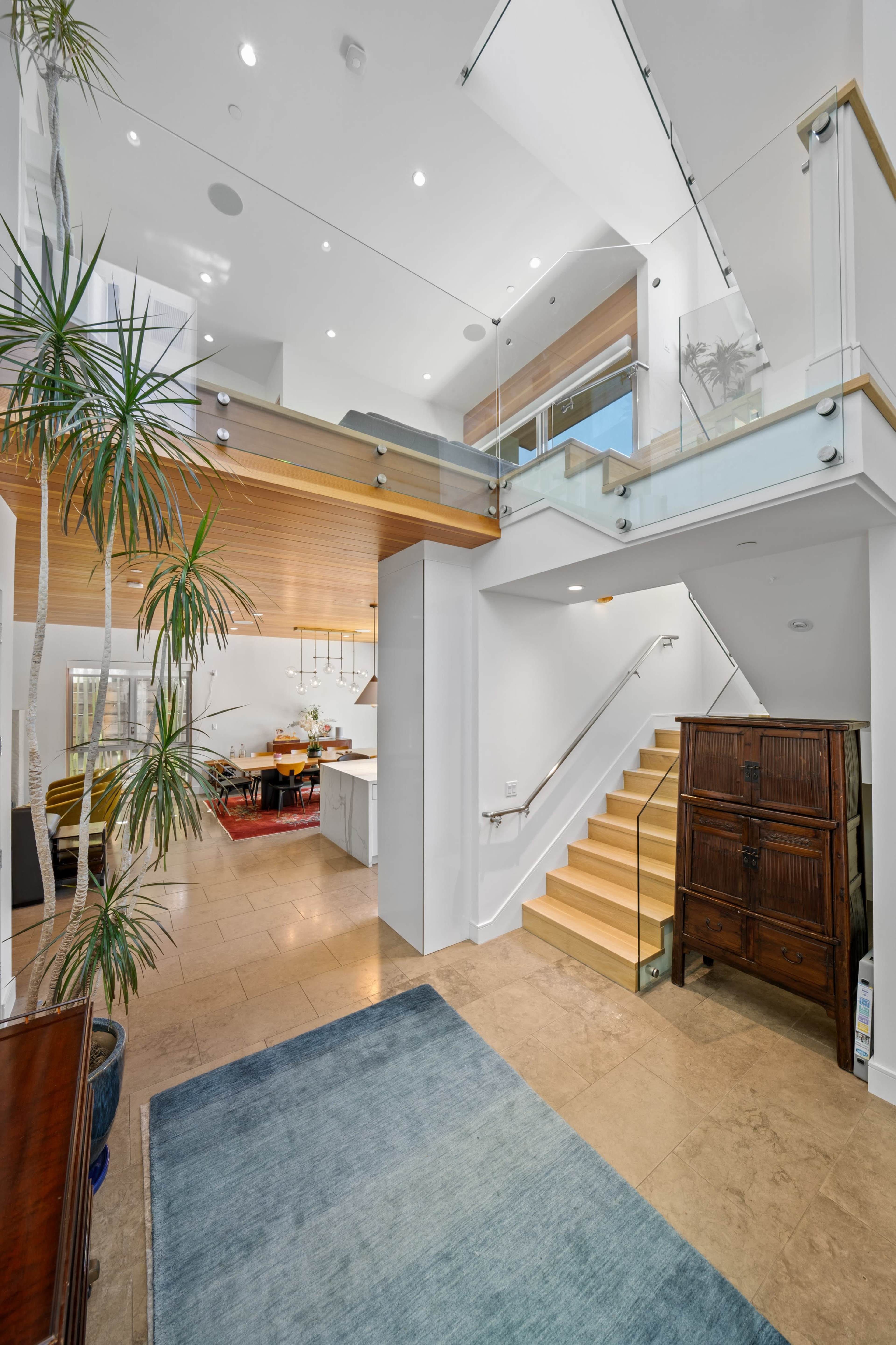 A modern foyer with a staircase leading to an upper level, featuring hardwood and glass elements, and a view of a dining area in the background.