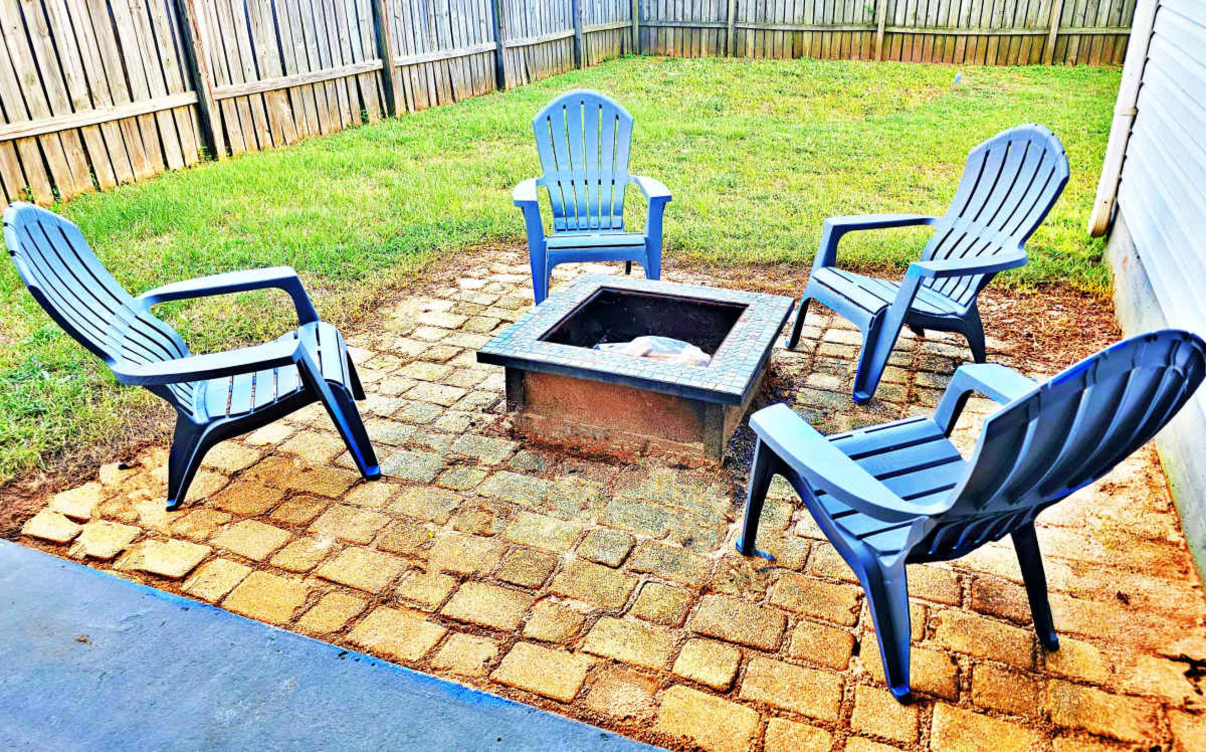 A fire pit surrounded by four blue chairs is situated on a stone patio in a grassy backyard.