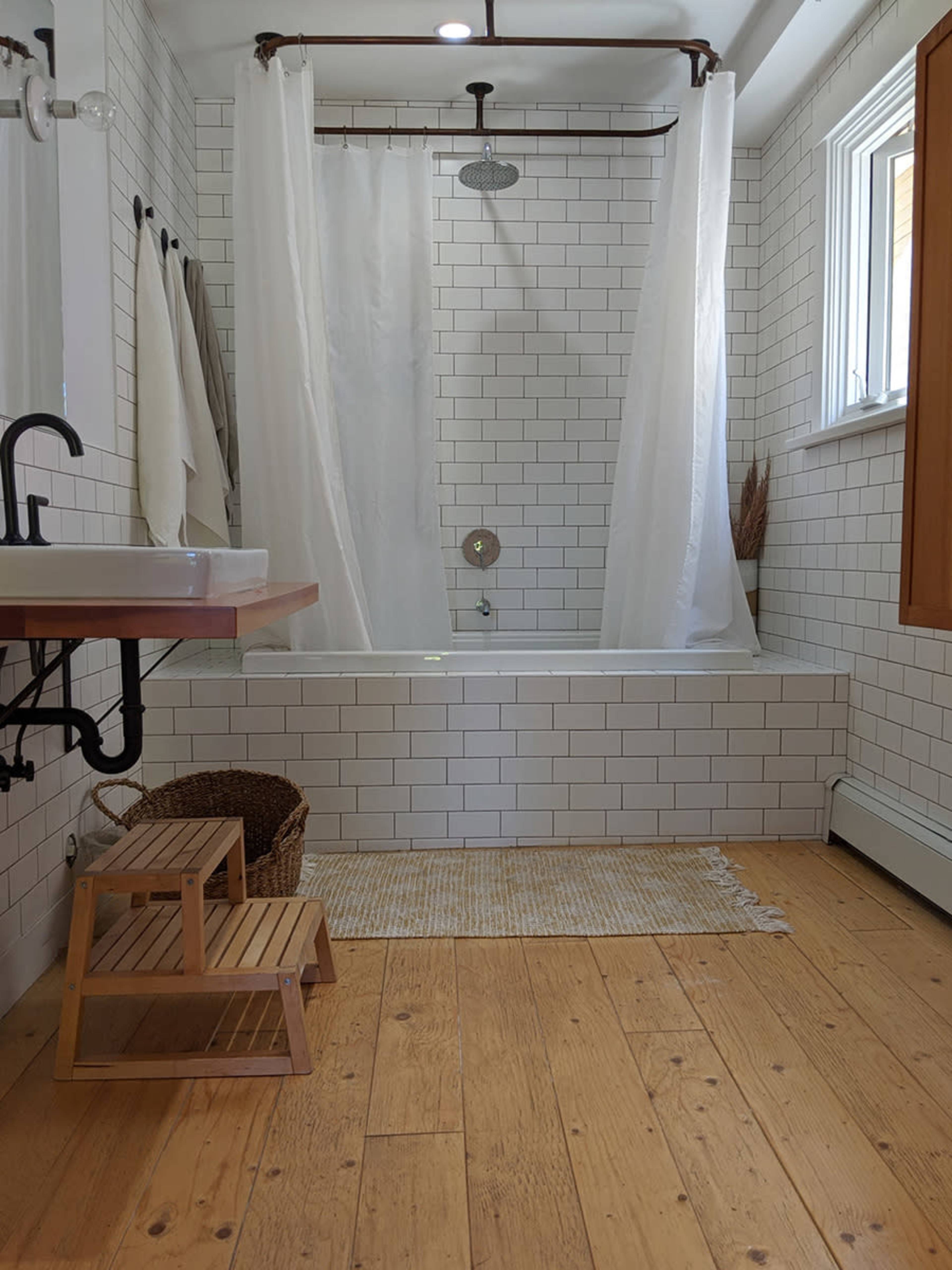 The image shows a modern bathroom featuring a bathtub with a shower curtain, a wooden step stool, and a washbasin on a wooden countertop among white tiled walls and a wooden floor.