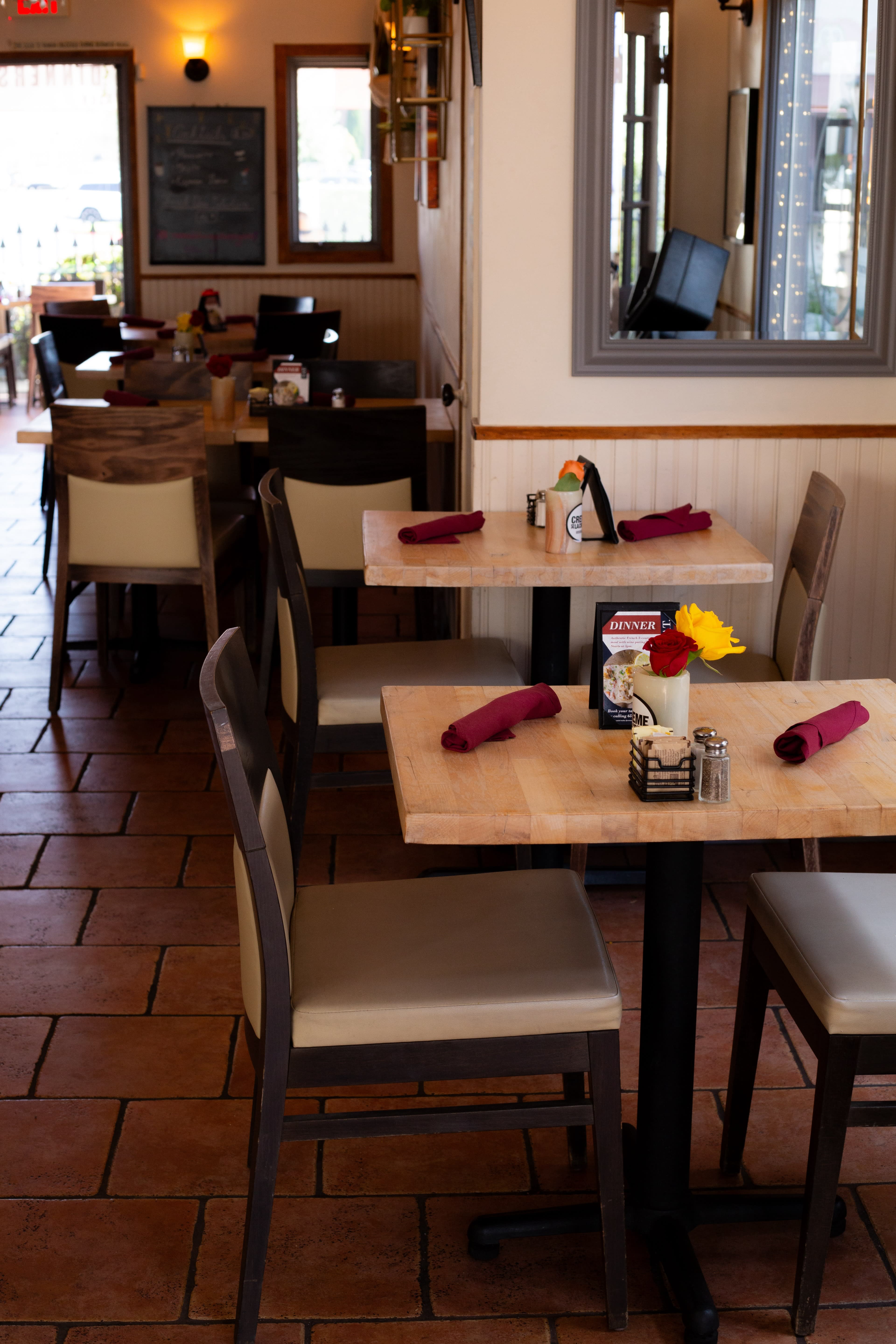 A restaurant interior features several wooden tables with chairs, set for dining with napkins and condiments arranged on each table.