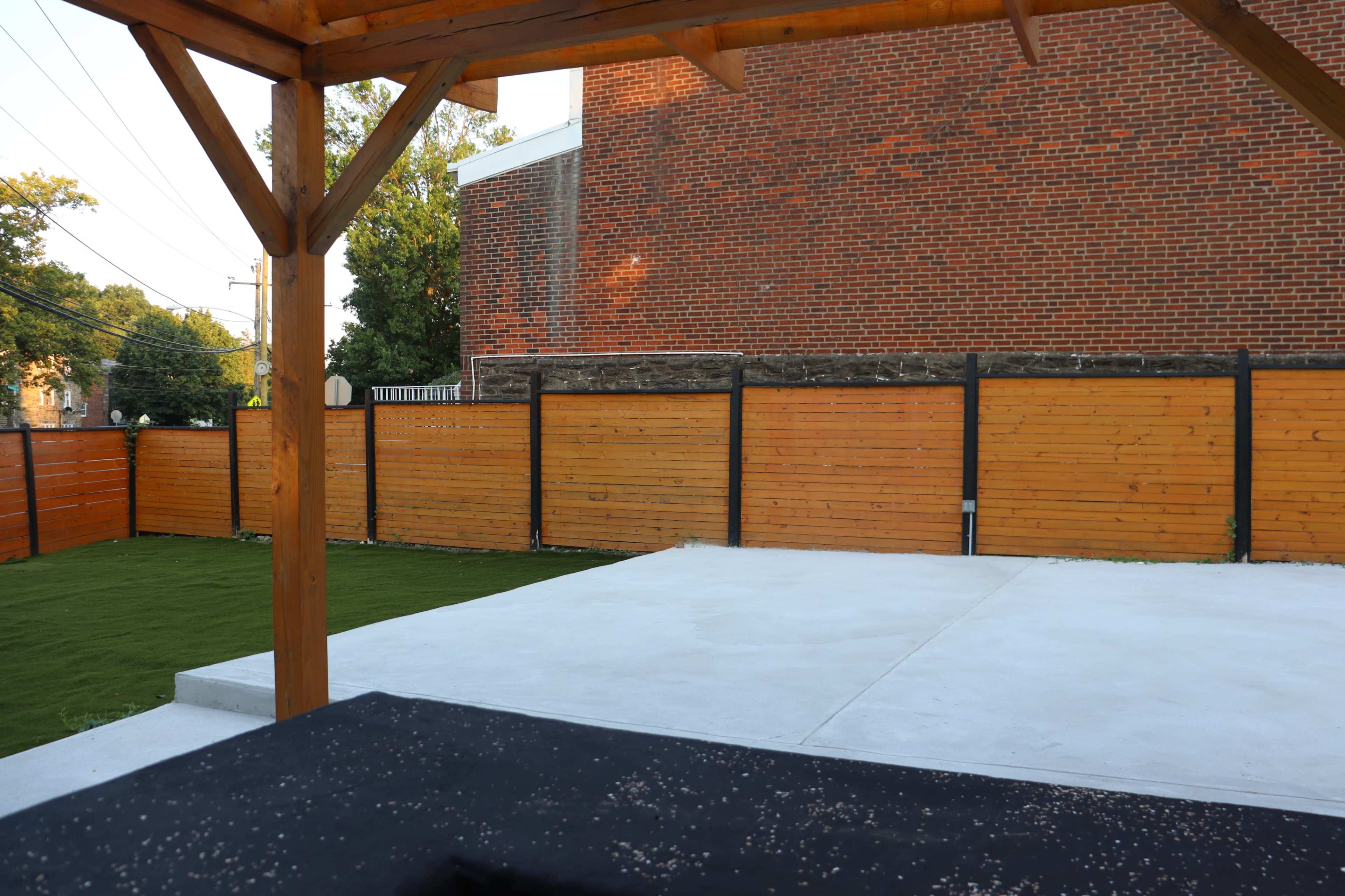 The image shows a patio area with a wooden pergola, concrete flooring, and a grassy section enclosed by wooden fencing and a brick building in the background.