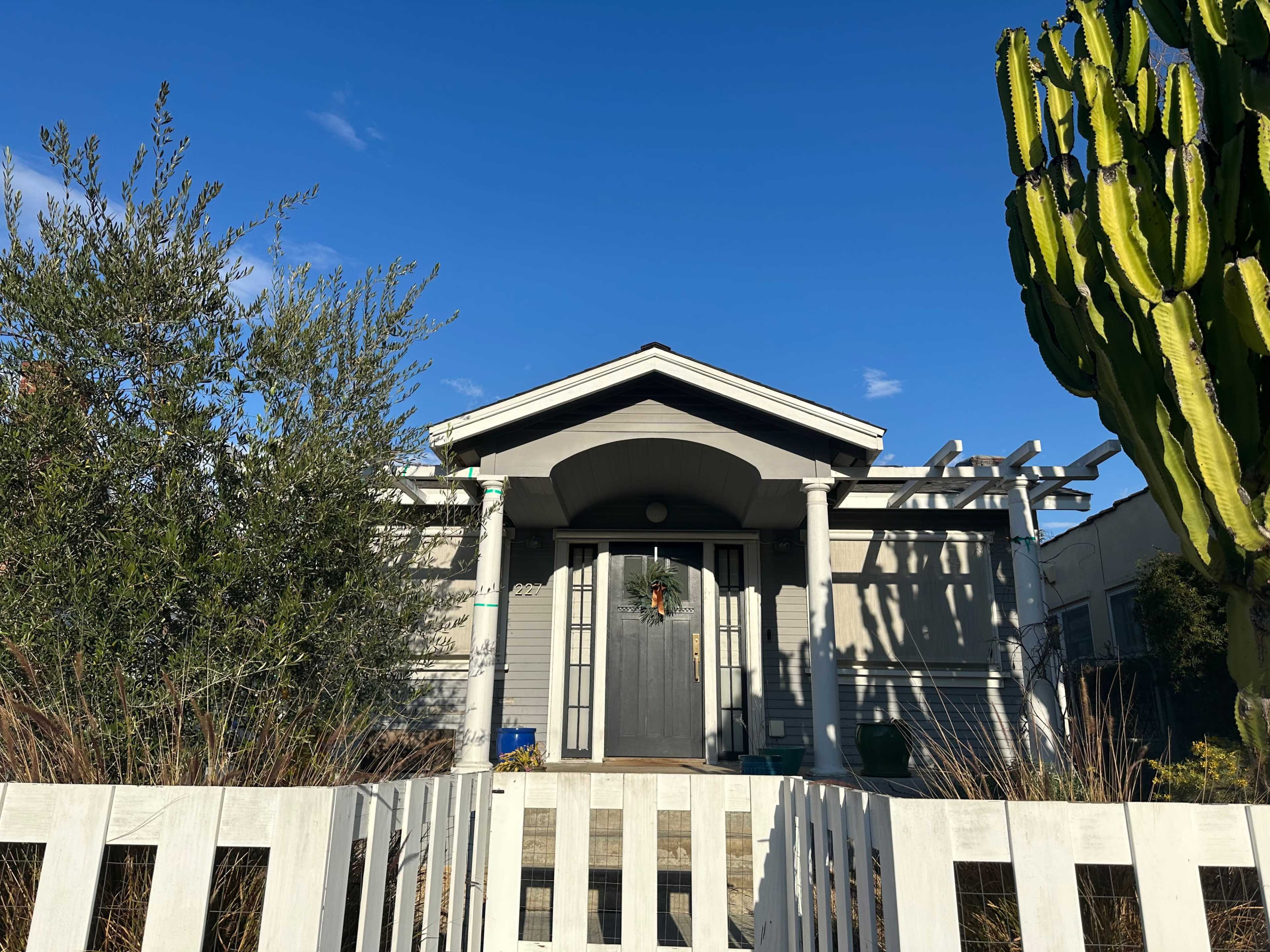 The image shows a gray house with a central front door, flanked by greenery and a tall cactus, under a clear blue sky.