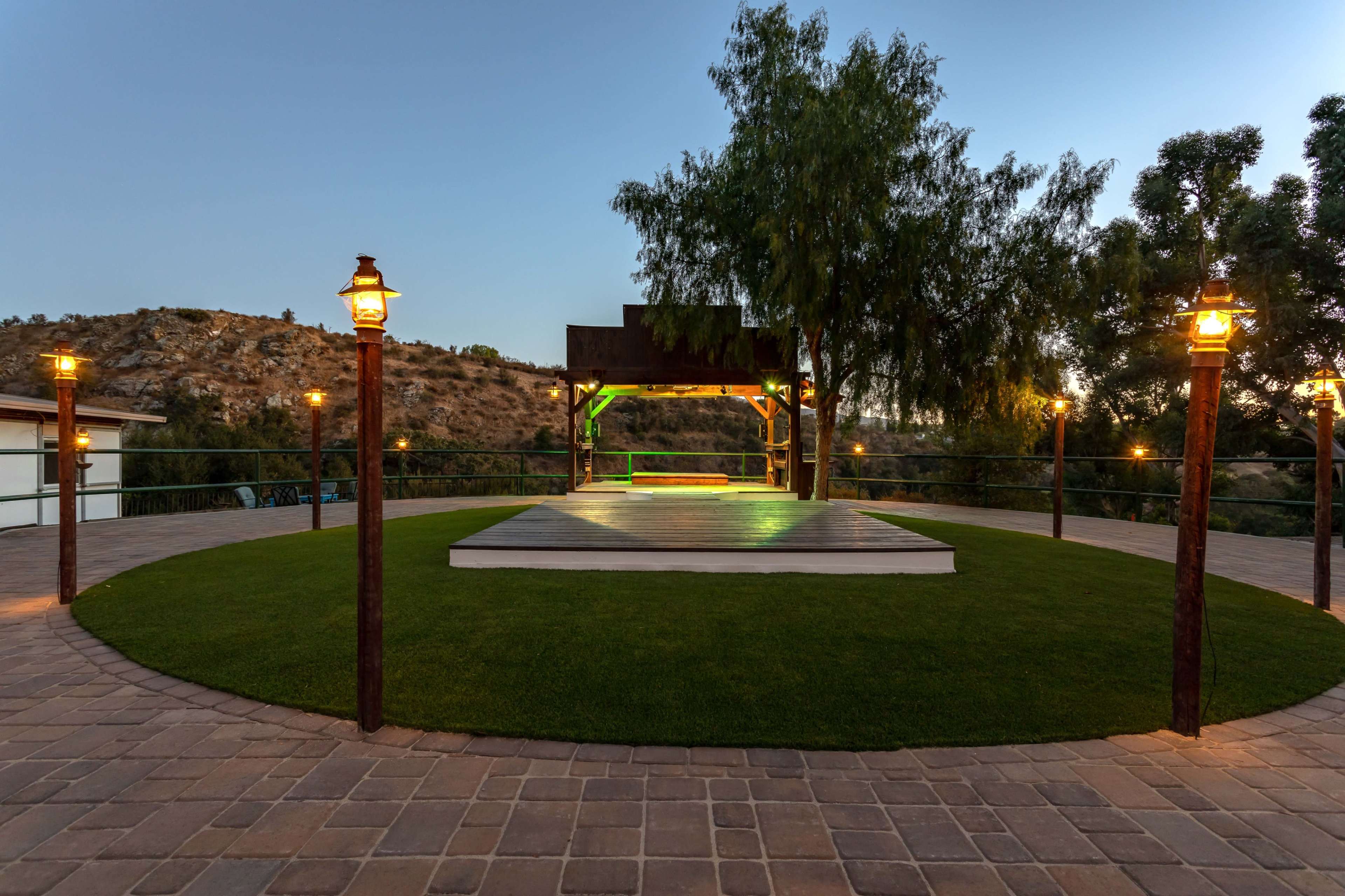 An outdoor stage area surrounded by low lighting and greenery, set against a hilly landscape at dusk.