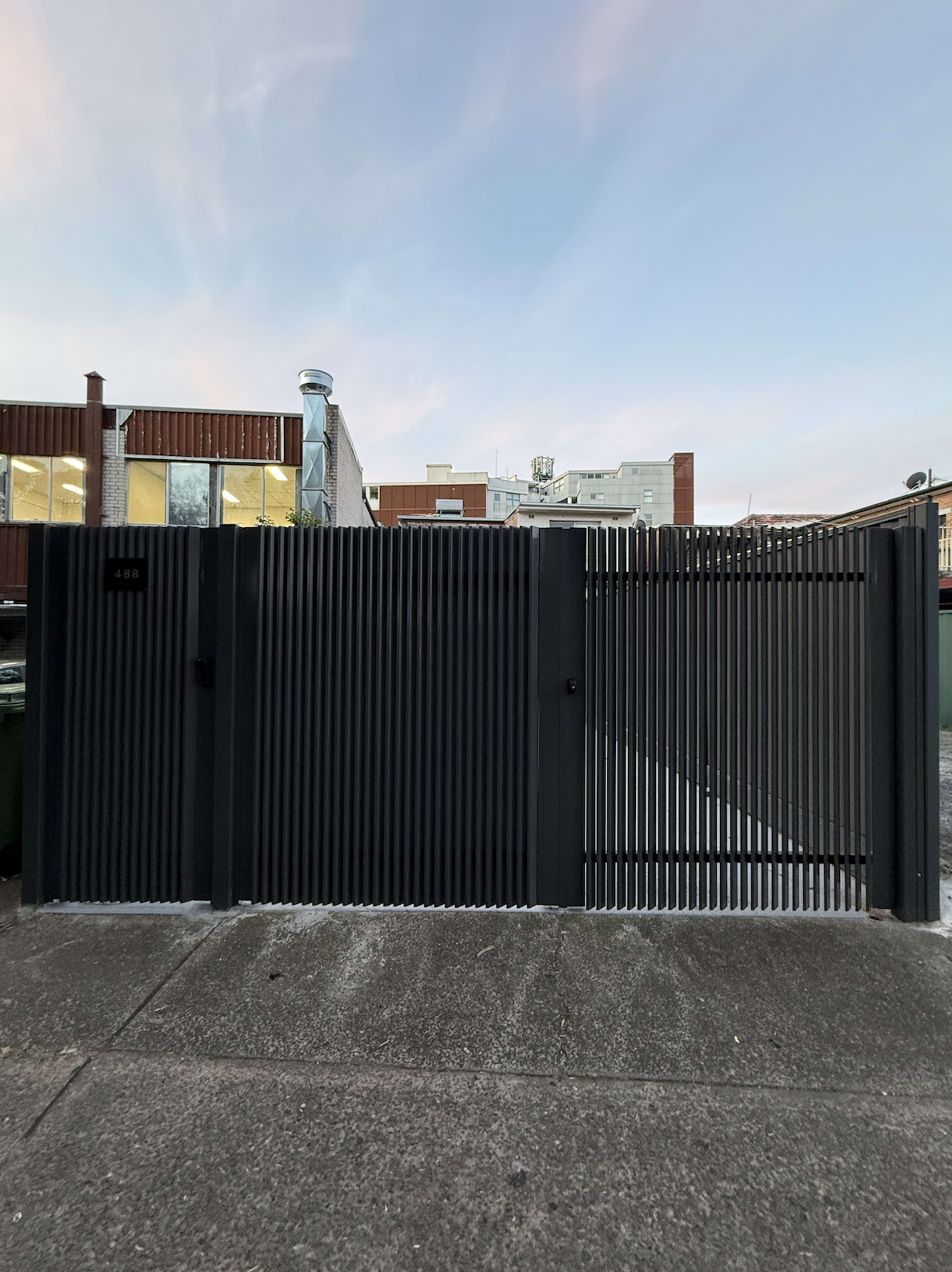 The image features a modern black metal gate with vertical slats, set against a backdrop of buildings and a blue sky.