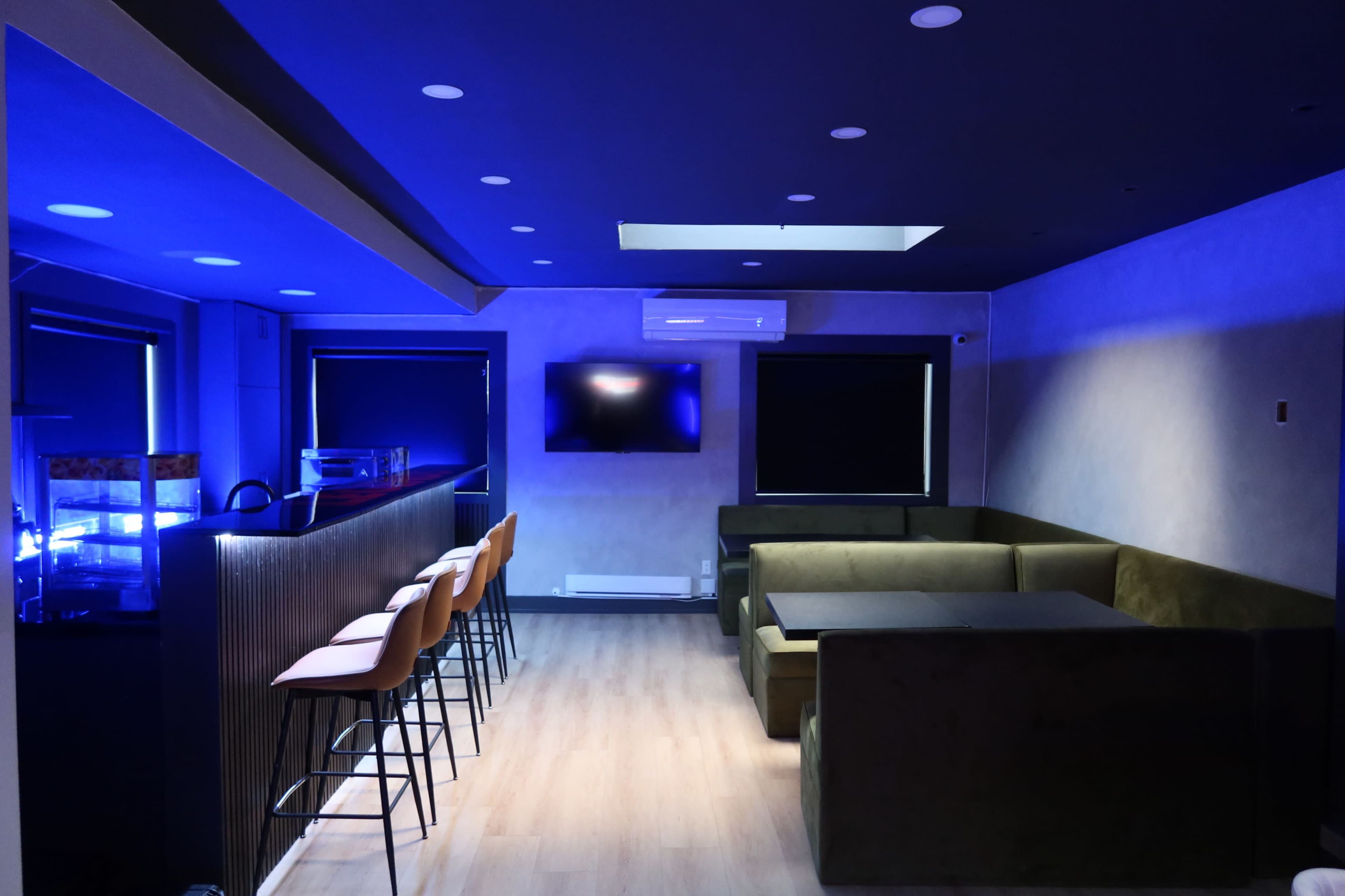 The image shows a modern lounge area with a bar, featuring blue lighting, a seating arrangement of a plush sofa, and several high stools.