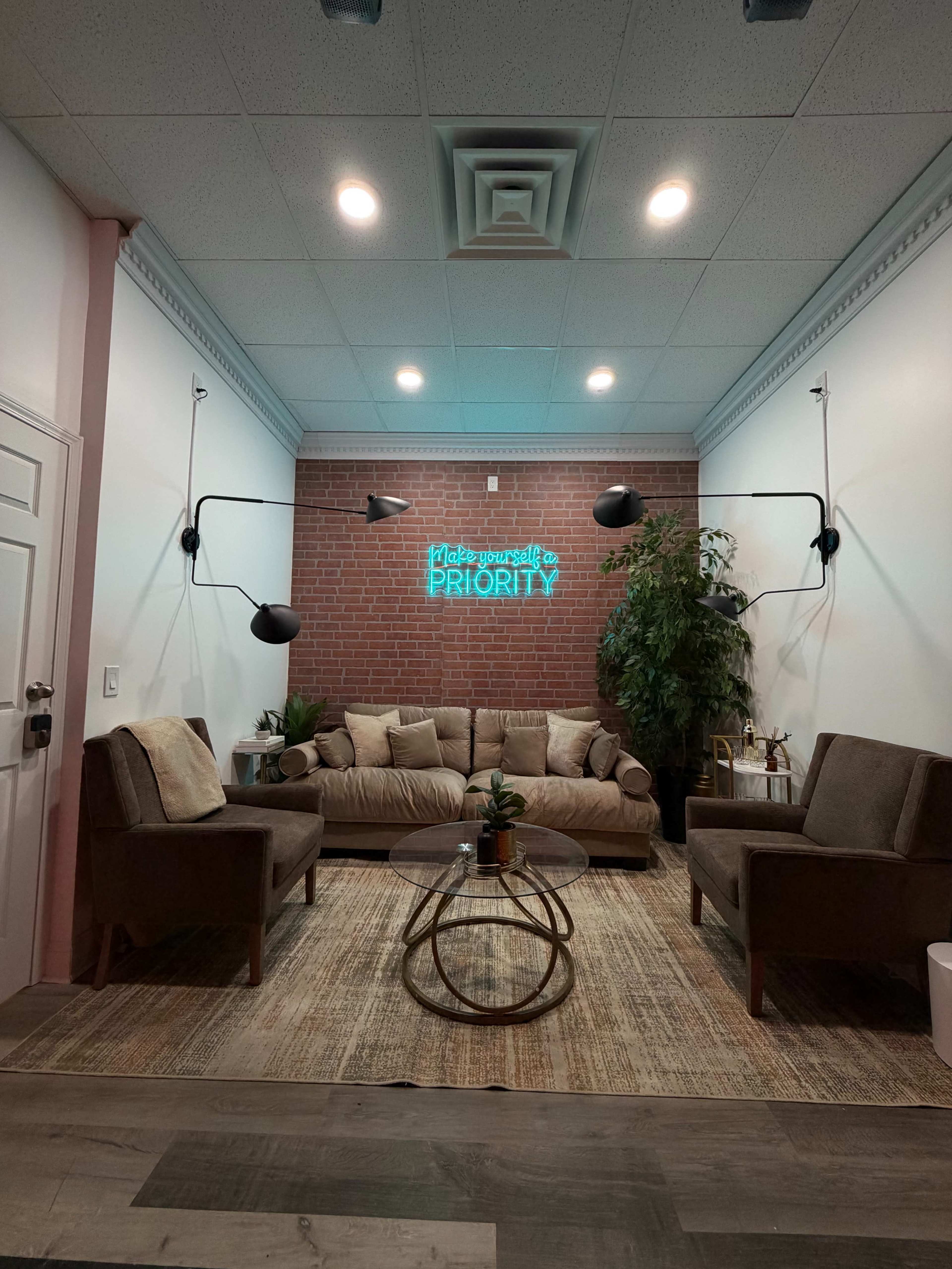 The image shows a cozy sitting area featuring a brown sofa, two armchairs, a round glass coffee table, and a plant, with a brick wall and neon sign in the background.