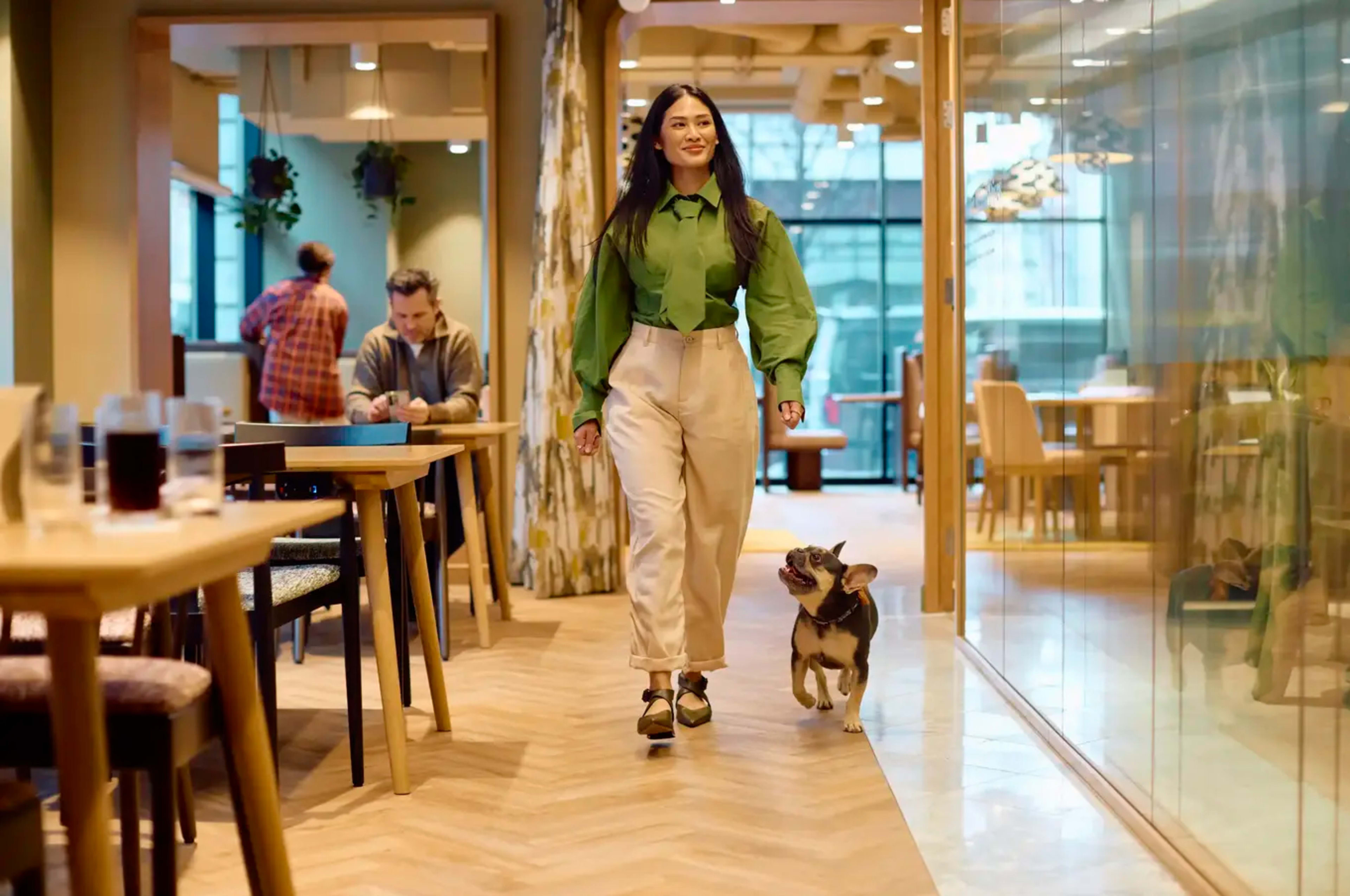A woman in a green top and beige pants walks alongside a small dog through a modern café with wooden furniture and glass walls.