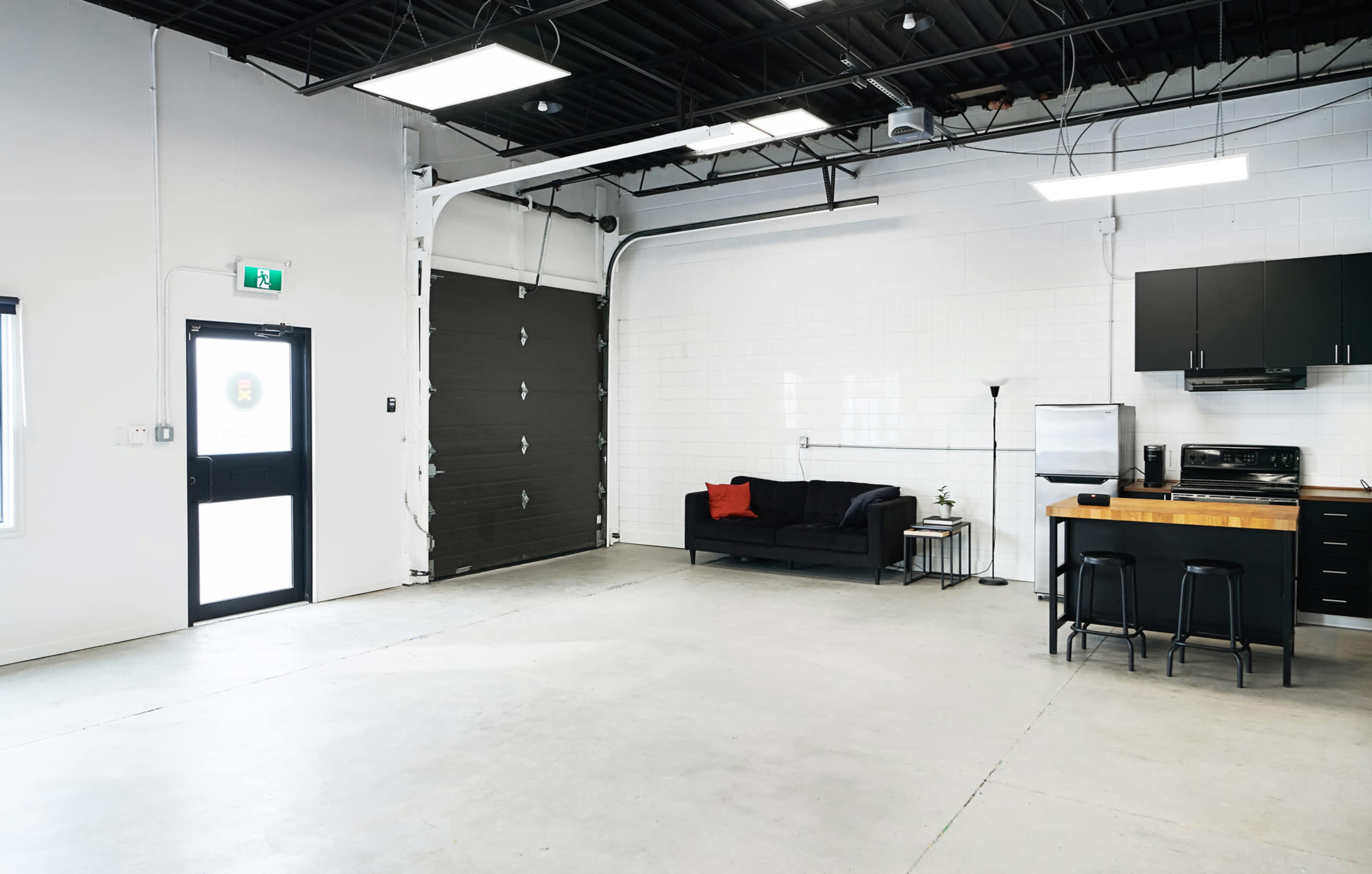 A spacious, modern room with a black couch, a small kitchen area, and a large garage door on one side.