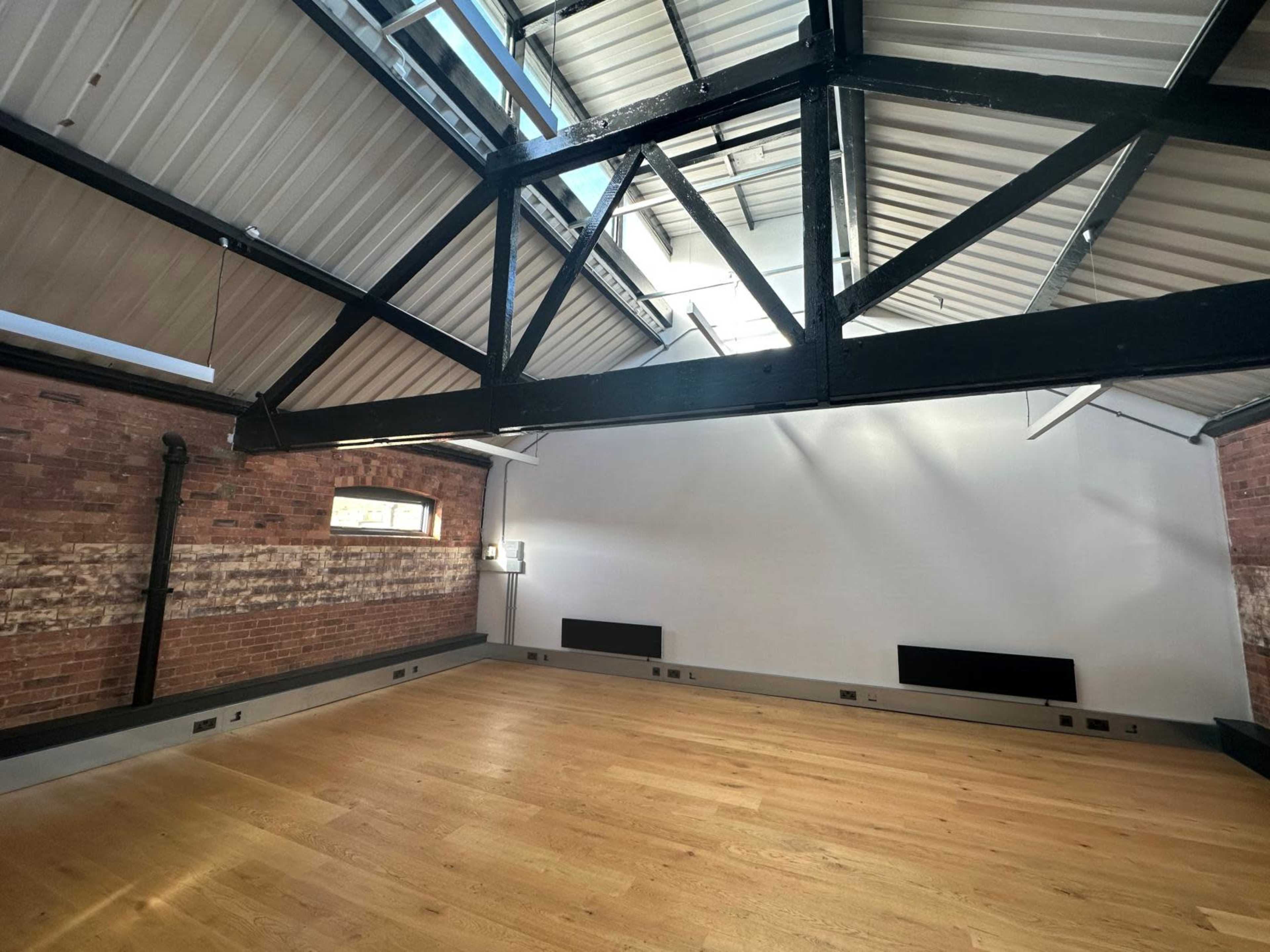 A spacious room with exposed brick walls, high ceilings, and a wooden floor, featuring large windows and black steel beams.
