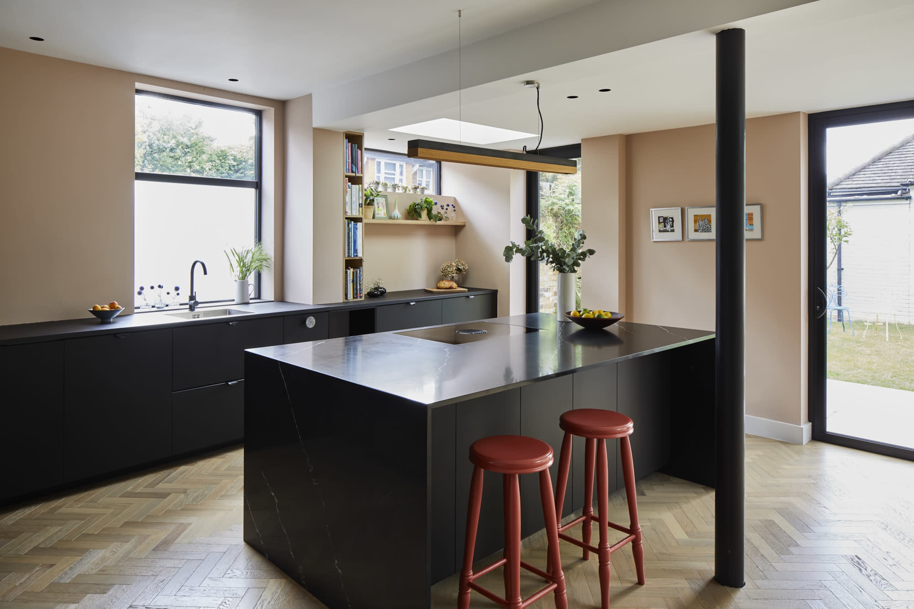 A modern kitchen features a dark cabinetry design, a central island with a metallic countertop, and red bar stools, with large windows allowing natural light to enter the space.