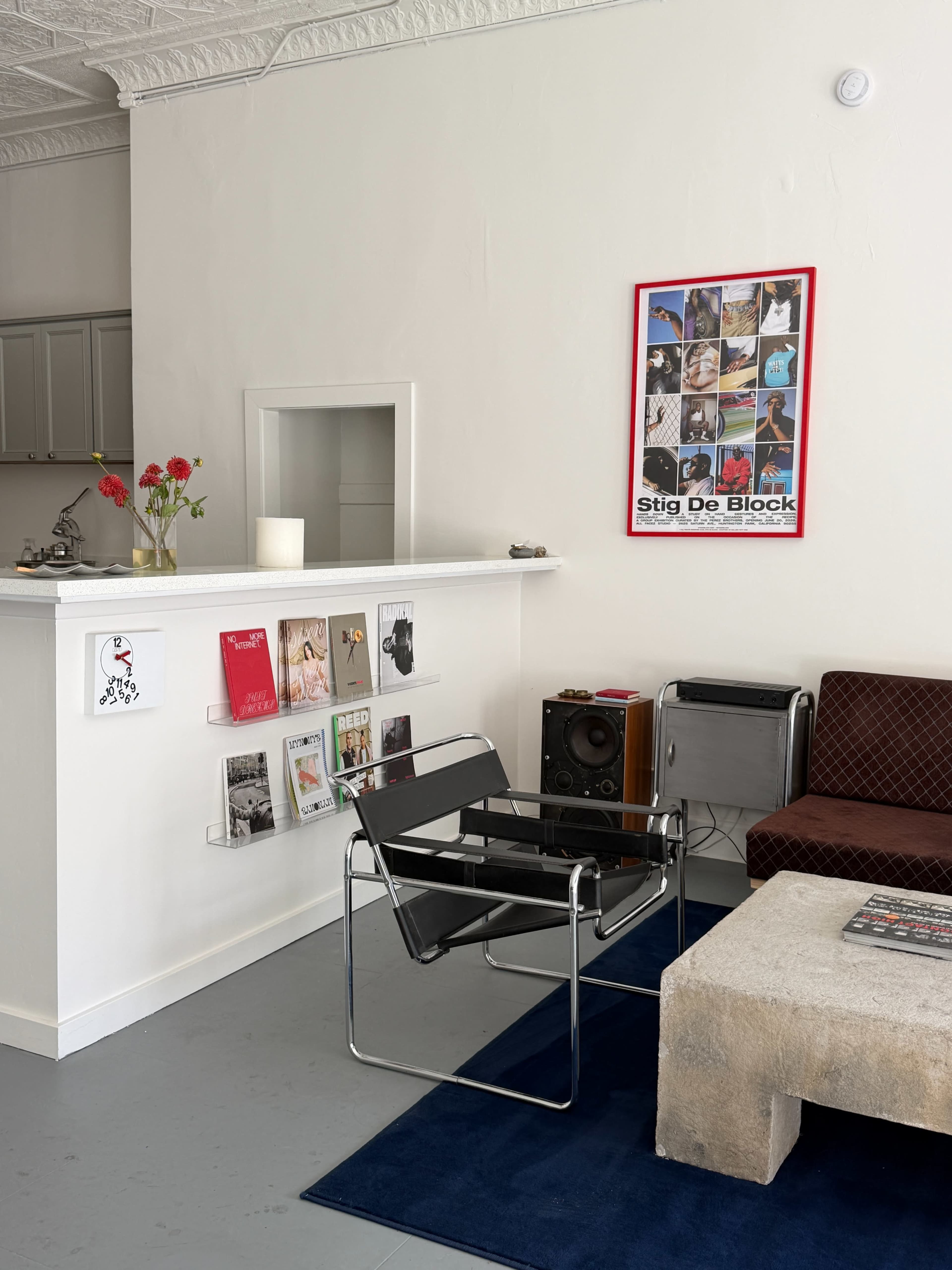 The image shows a minimalist interior with a black chair, a brown couch, a concrete coffee table, a small flower arrangement, and a wall displaying framed posters and magazines.