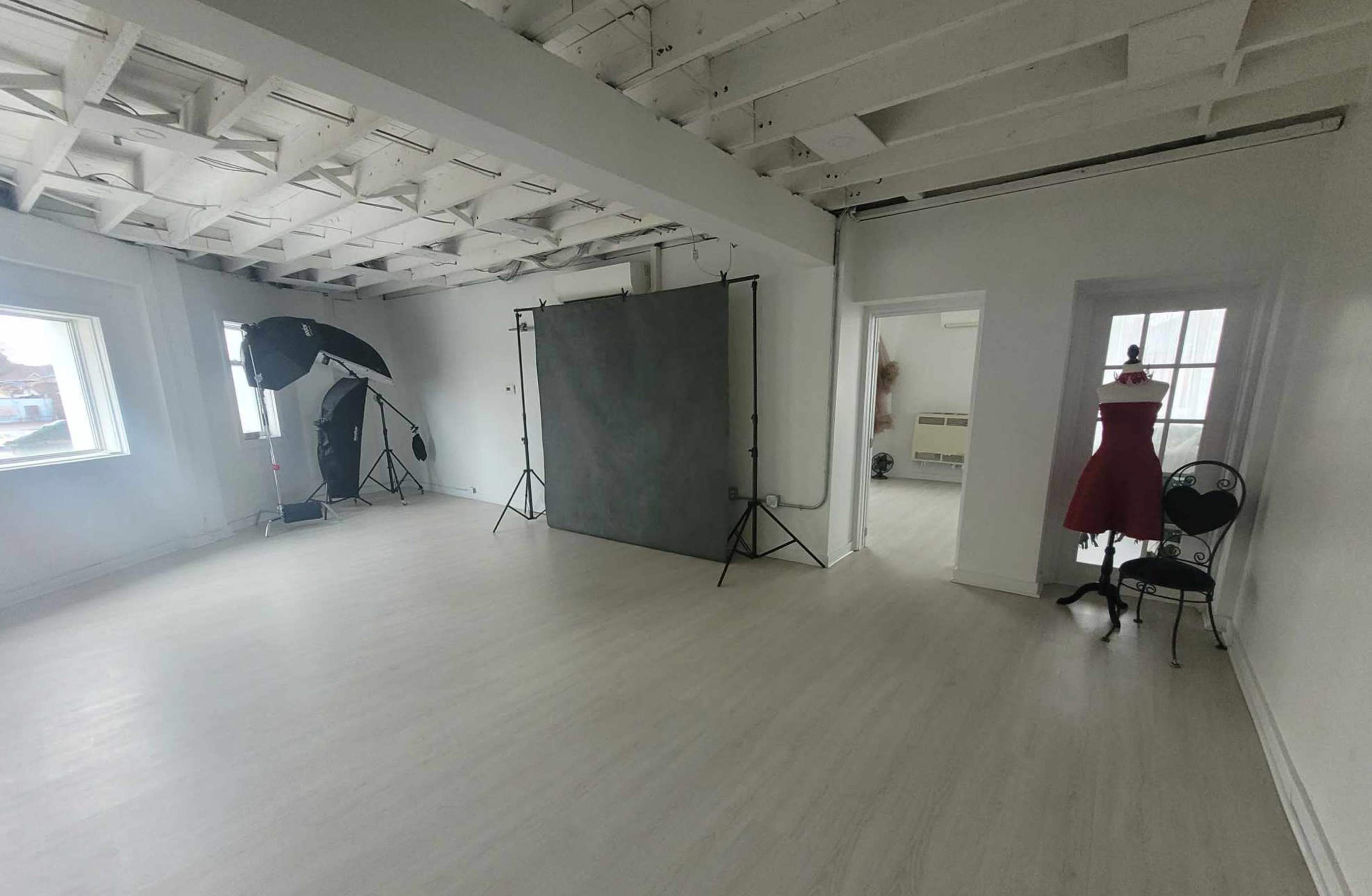 A bright, empty studio space with white walls, a light wooden floor, and a backdrop setup for photography, accompanied by a dress form in a corner.