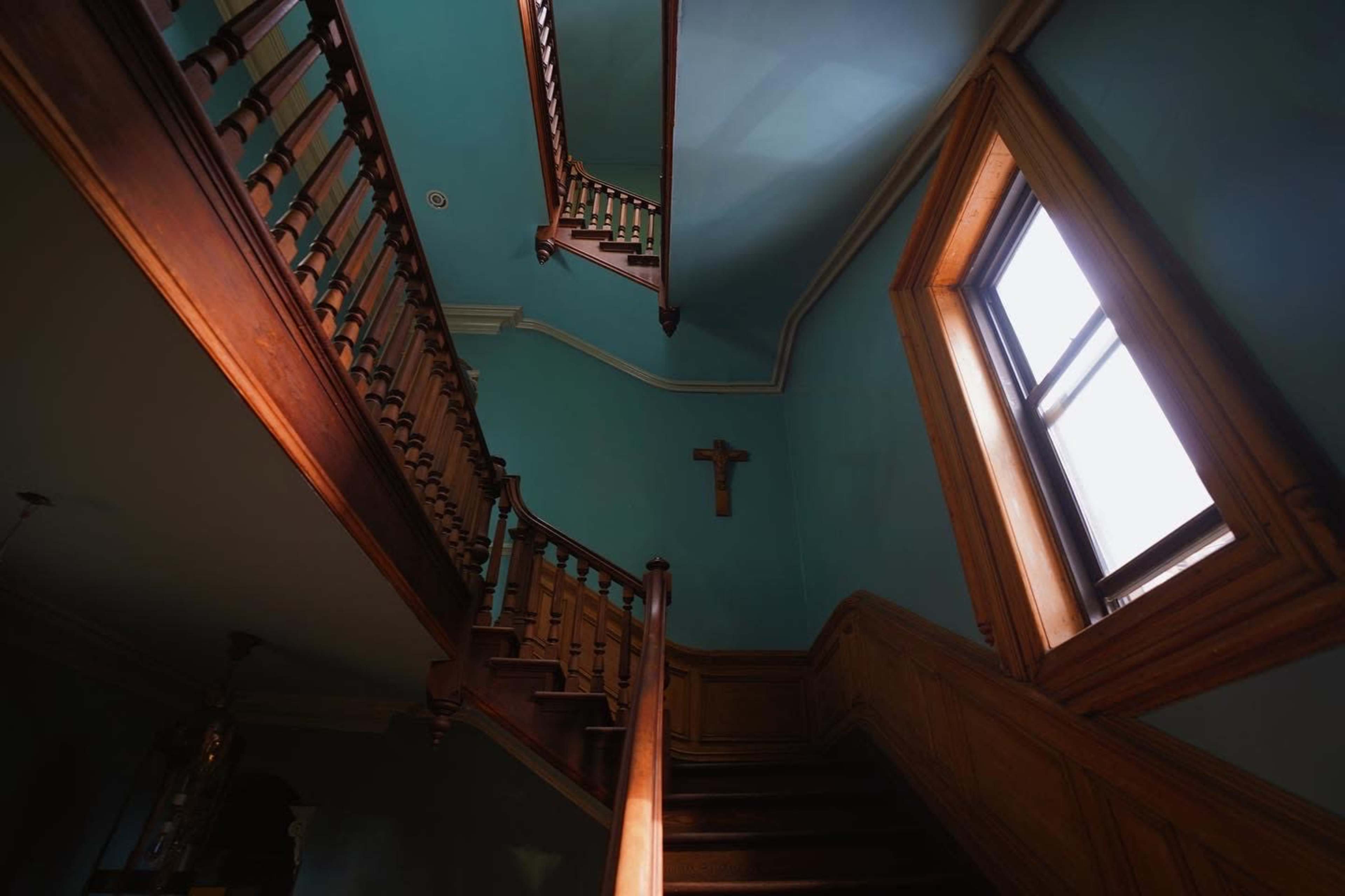 A wooden staircase ascends alongside turquoise walls, with a window and a cross visible on the wall above.