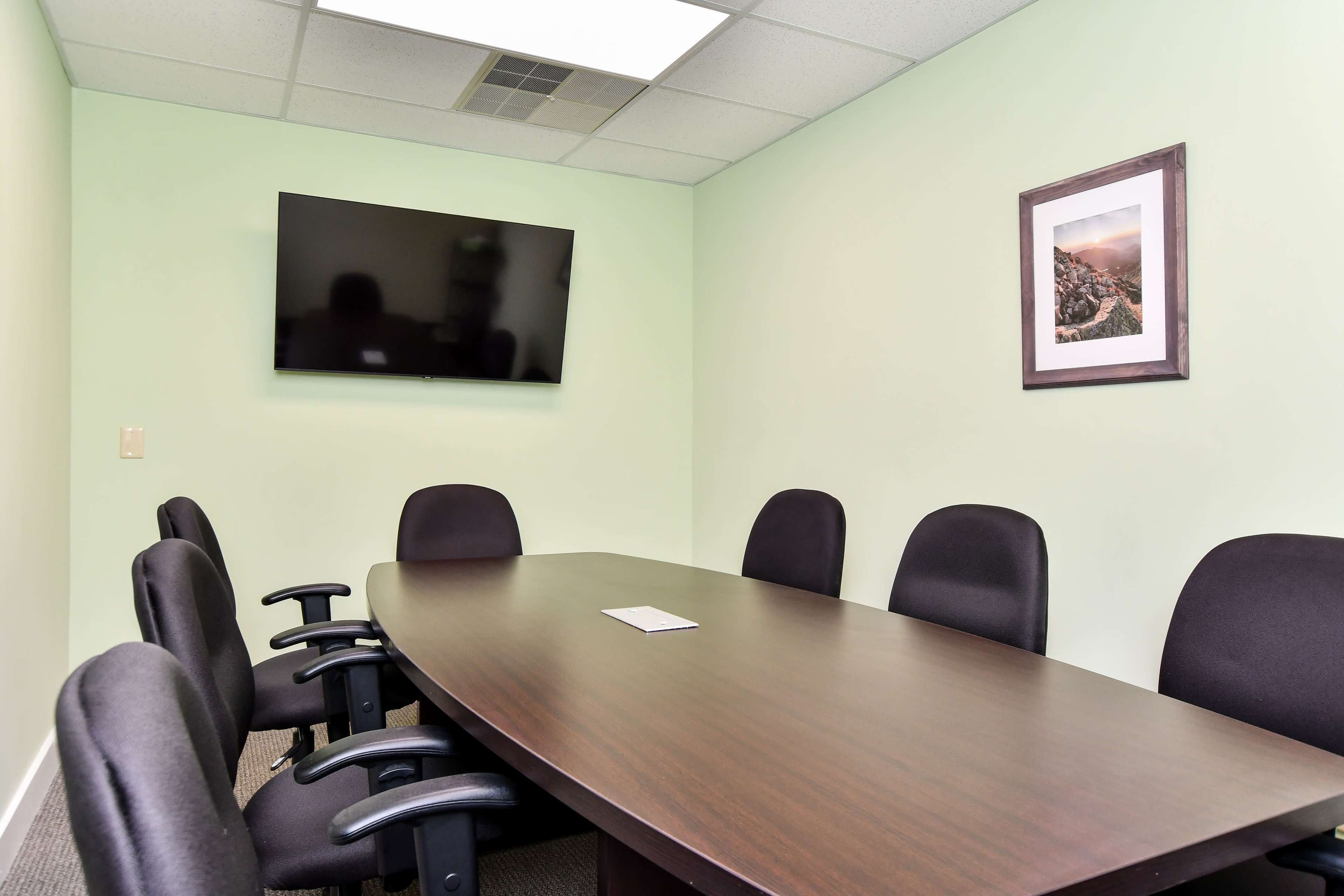 A conference room features a large wooden table surrounded by six black chairs, with a mounted television and a framed photo on the green wall.