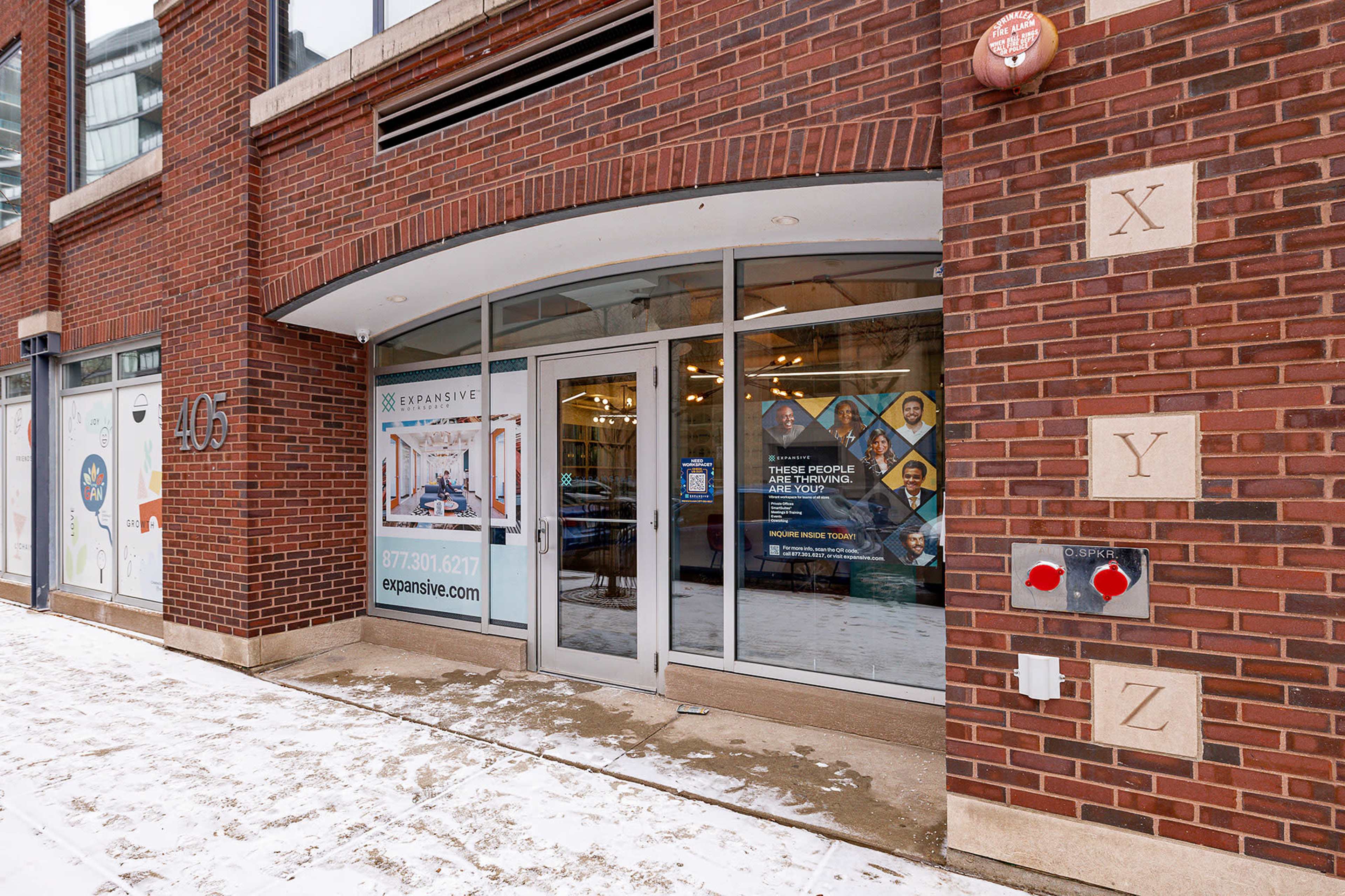 The image shows a modern brick building entrance with large glass doors and signs for businesses and directories, surrounded by light snow on the ground.