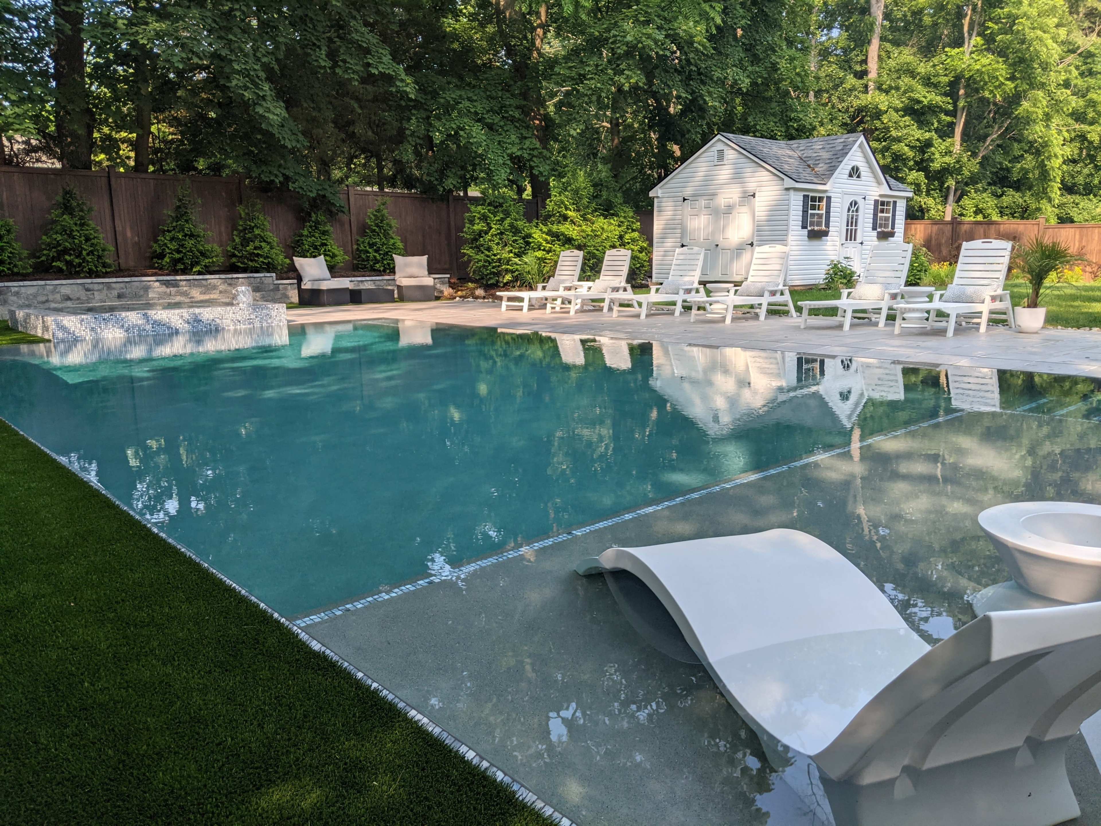 A clear swimming pool is surrounded by white lounge chairs and a small shed amidst lush greenery.