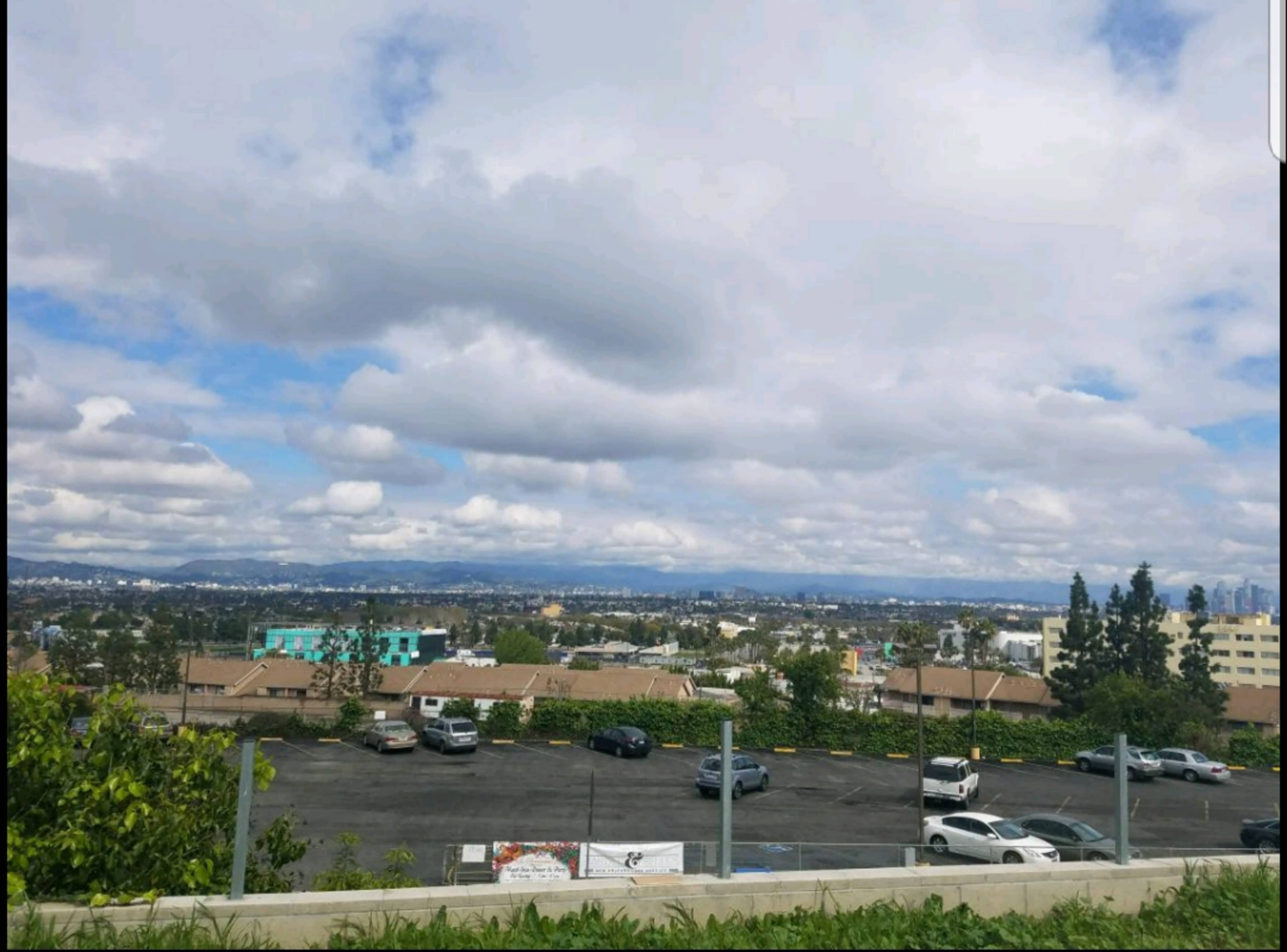 A wide view of a suburban area under a partly cloudy sky, with parked cars and distant mountains in the background.