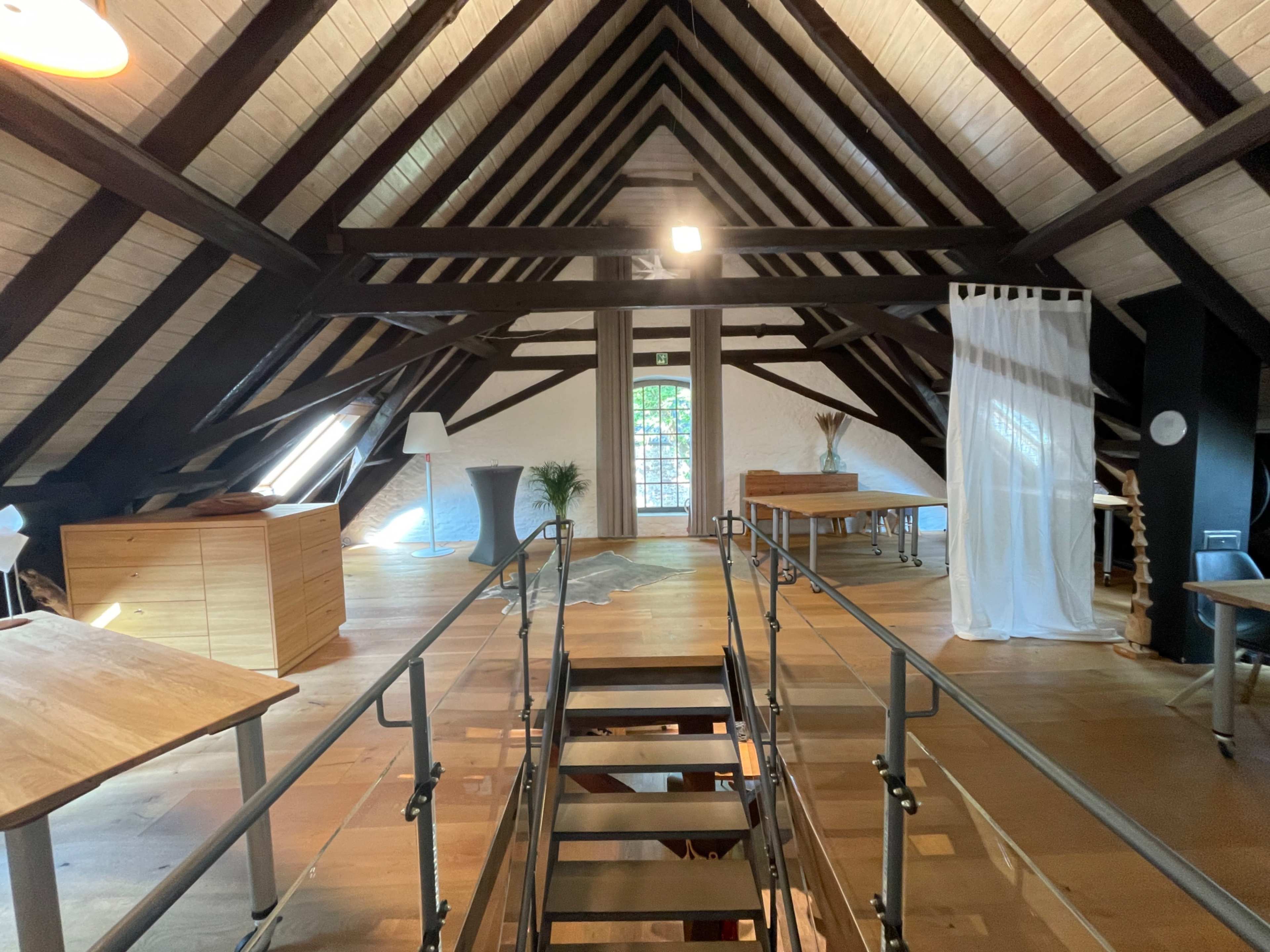 The image shows a spacious, well-lit attic with exposed wooden beams, a metal staircase, and several wooden tables.