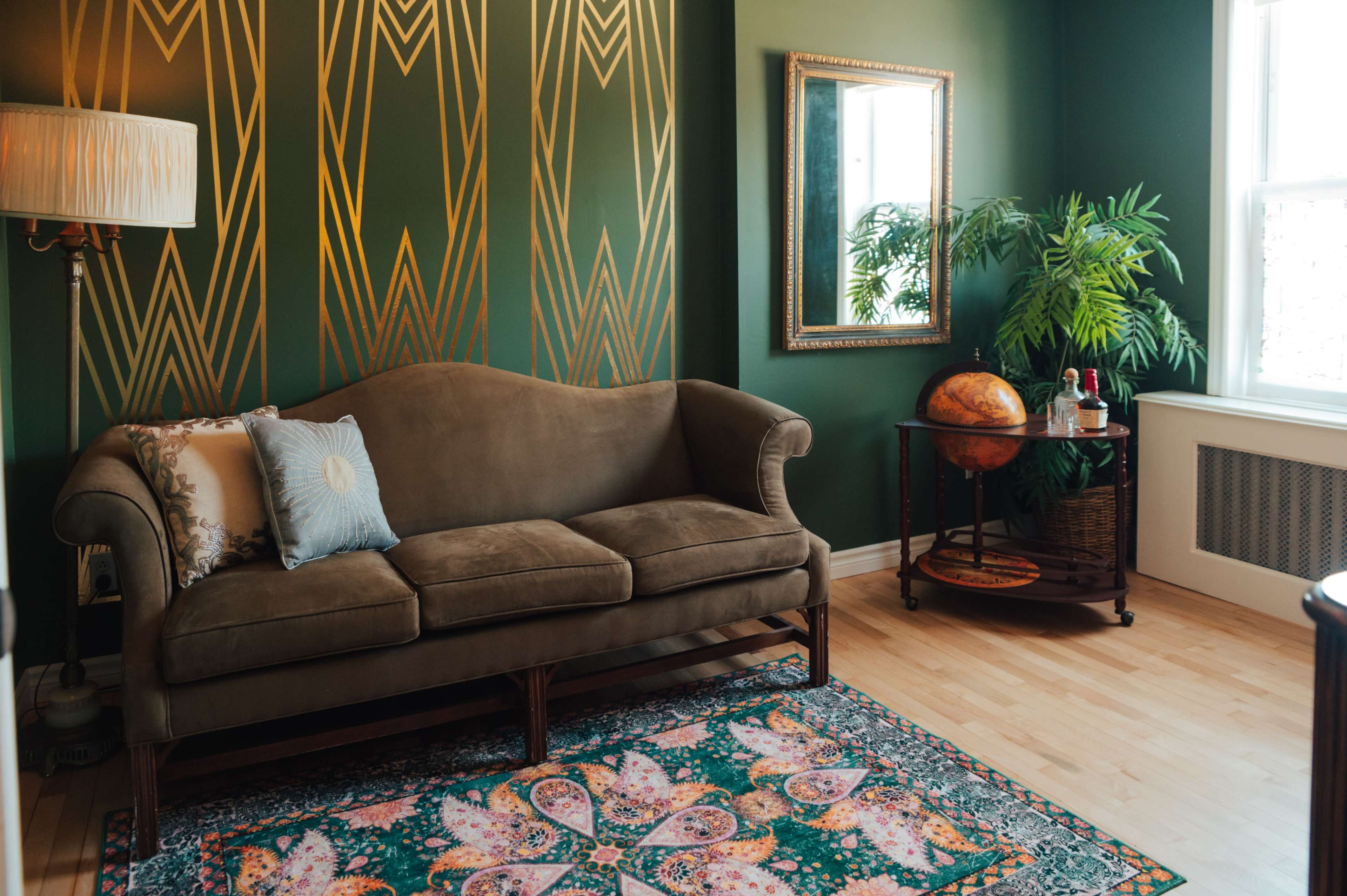 The image shows a living room featuring a brown sofa, a decorative area rug, a potted plant, a globe on a cart, and a mirror on a green accent wall with gold geometric patterns.