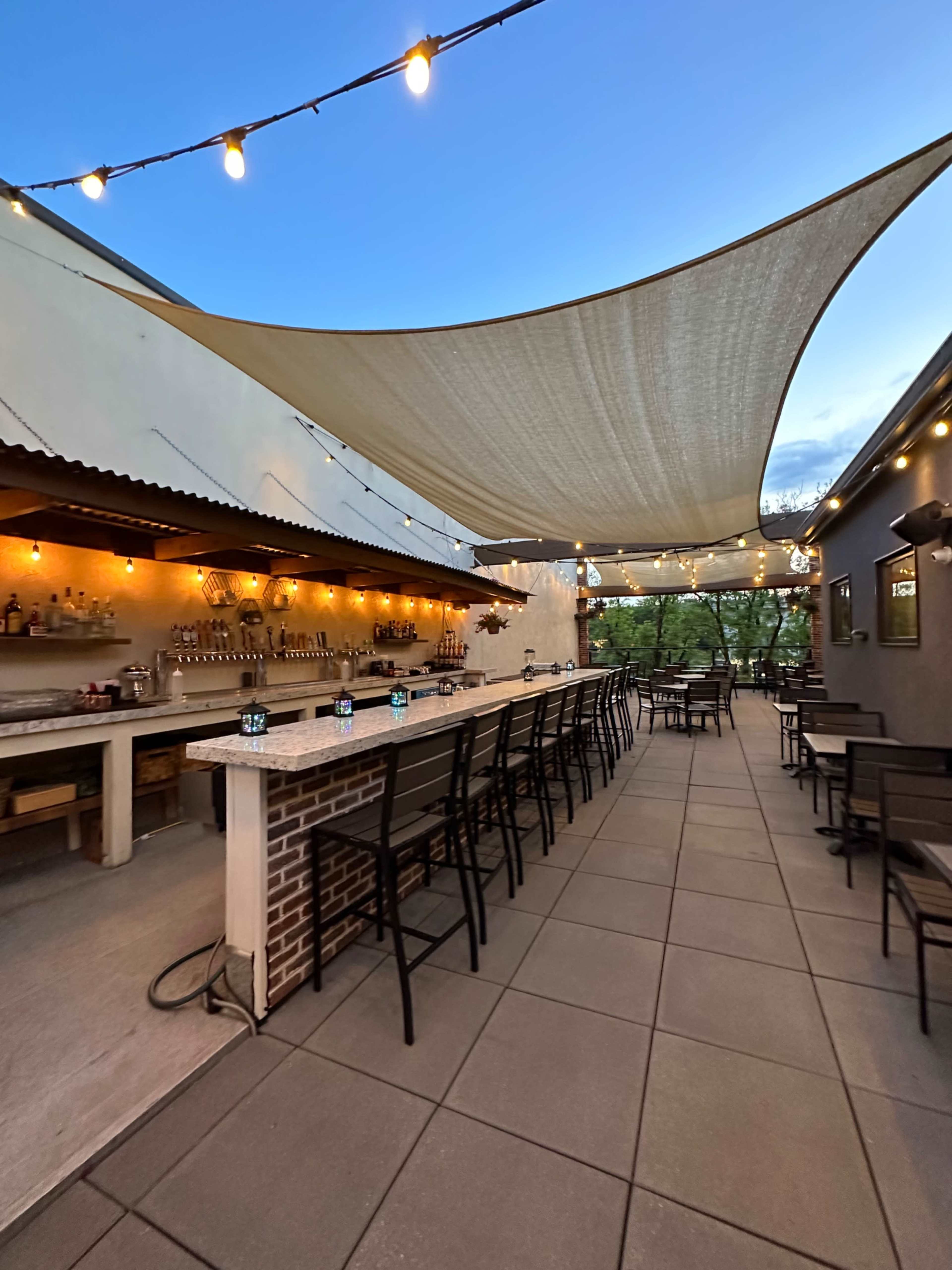 The image shows a spacious outdoor bar area with a long countertop, rows of chairs, and string lights overhead, set against a twilight sky.