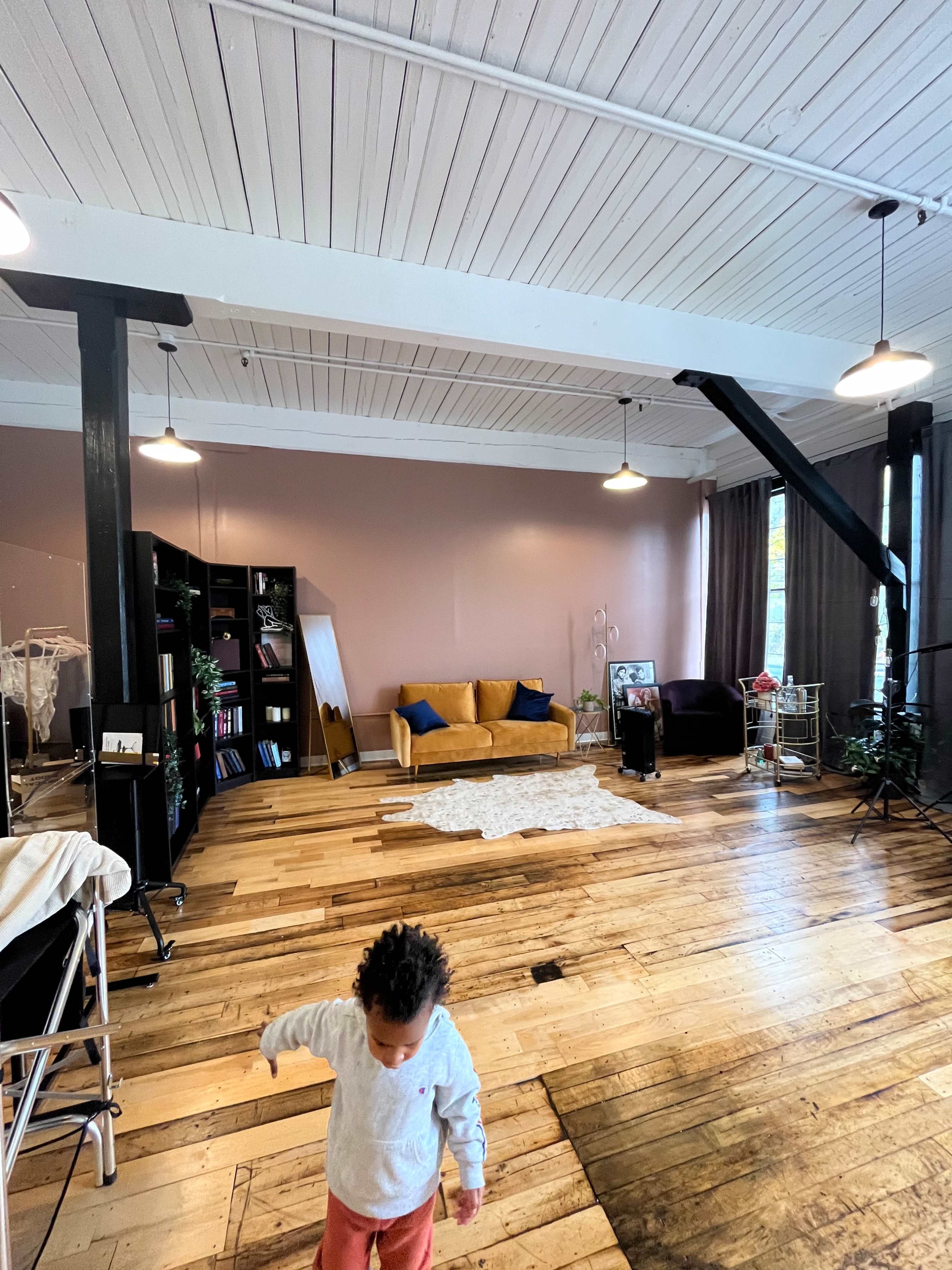 A child walks across a spacious room with wooden floors, featuring a couch, a bookshelf, and large windows.