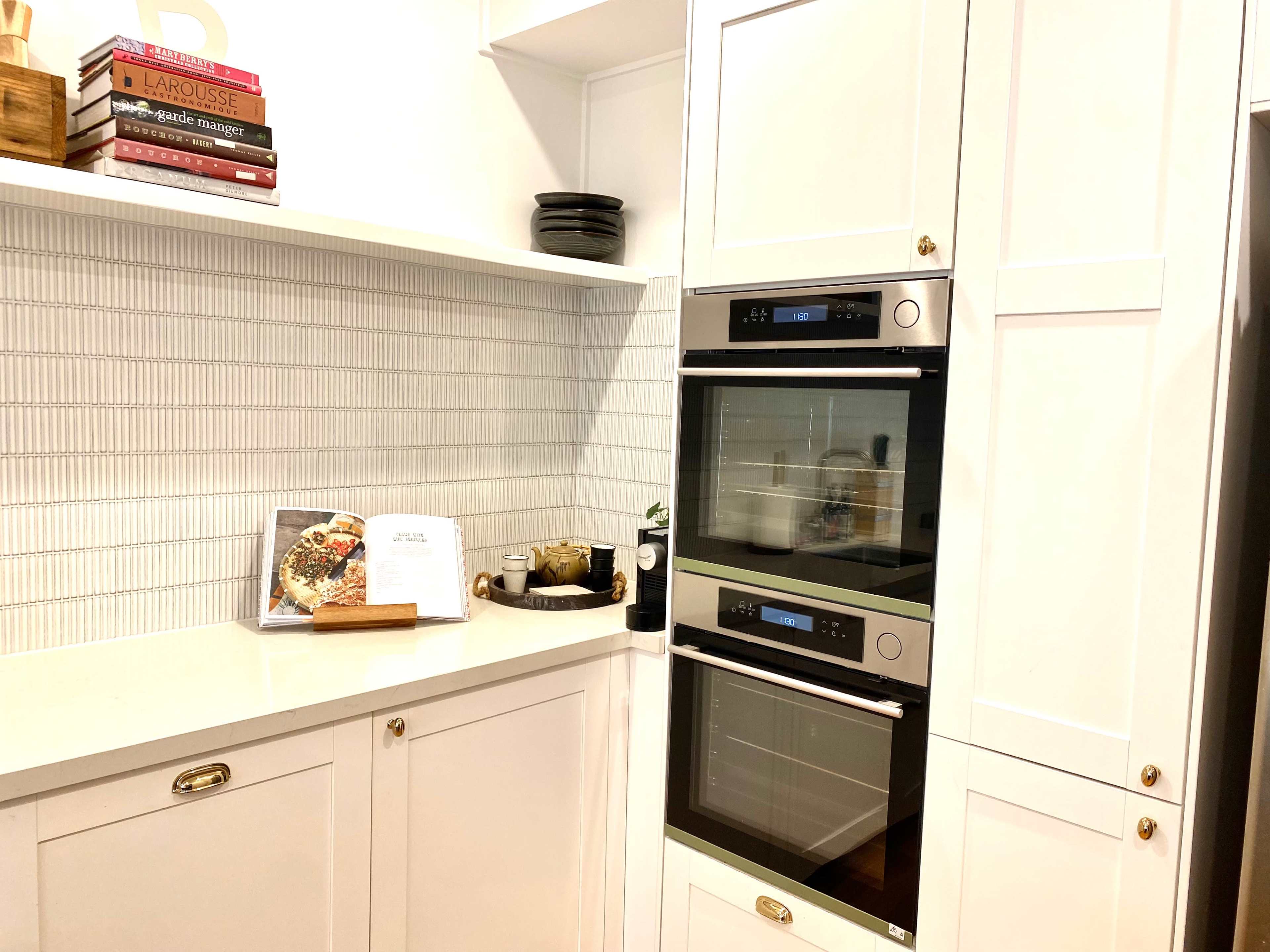 A modern kitchen corner featuring two stacked ovens, a white tiled backsplash, and neatly arranged kitchen accessories on a countertop.