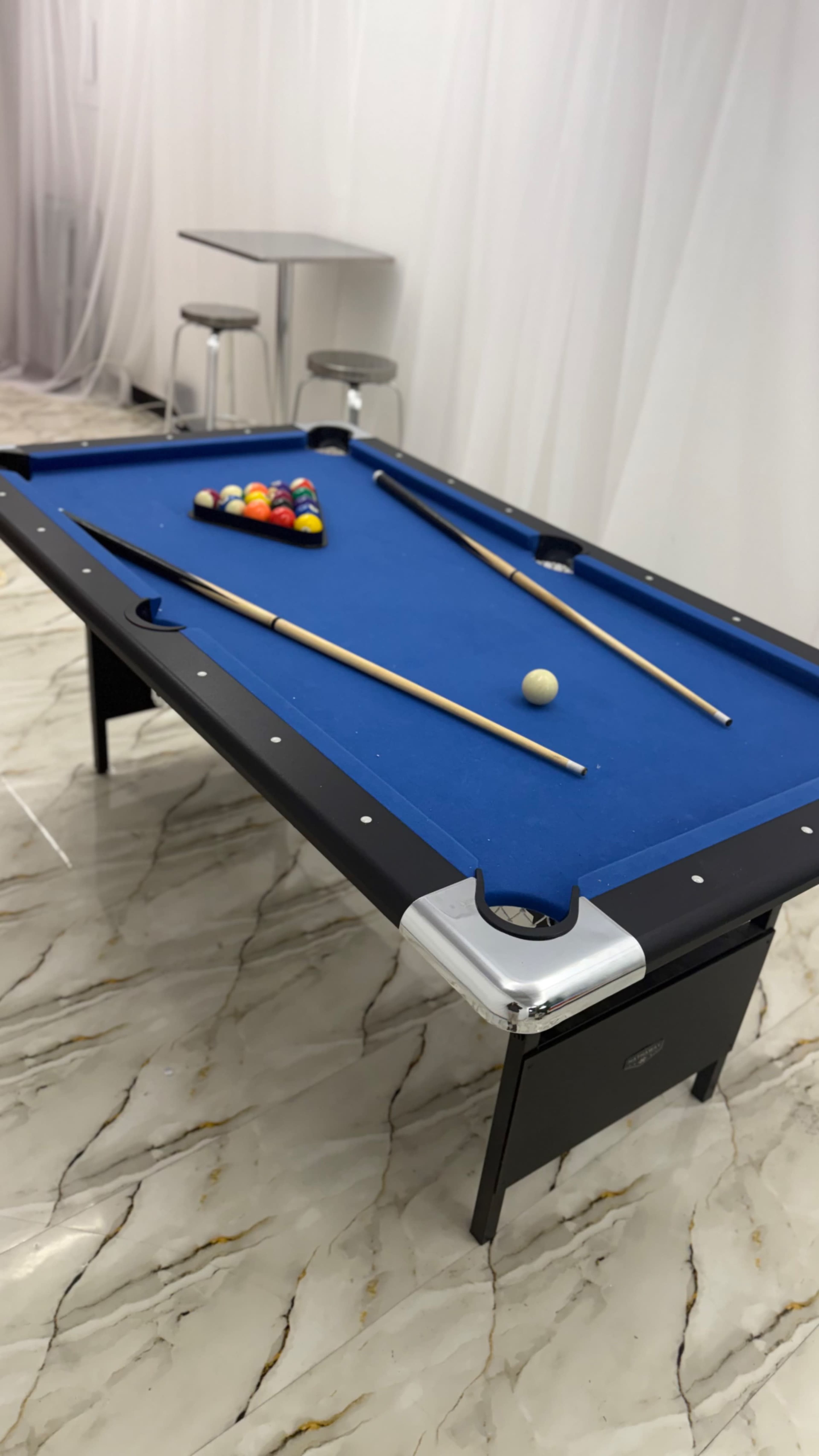 A blue felt pool table is set up with a triangle of colored balls at one end and two cue sticks positioned on the table.