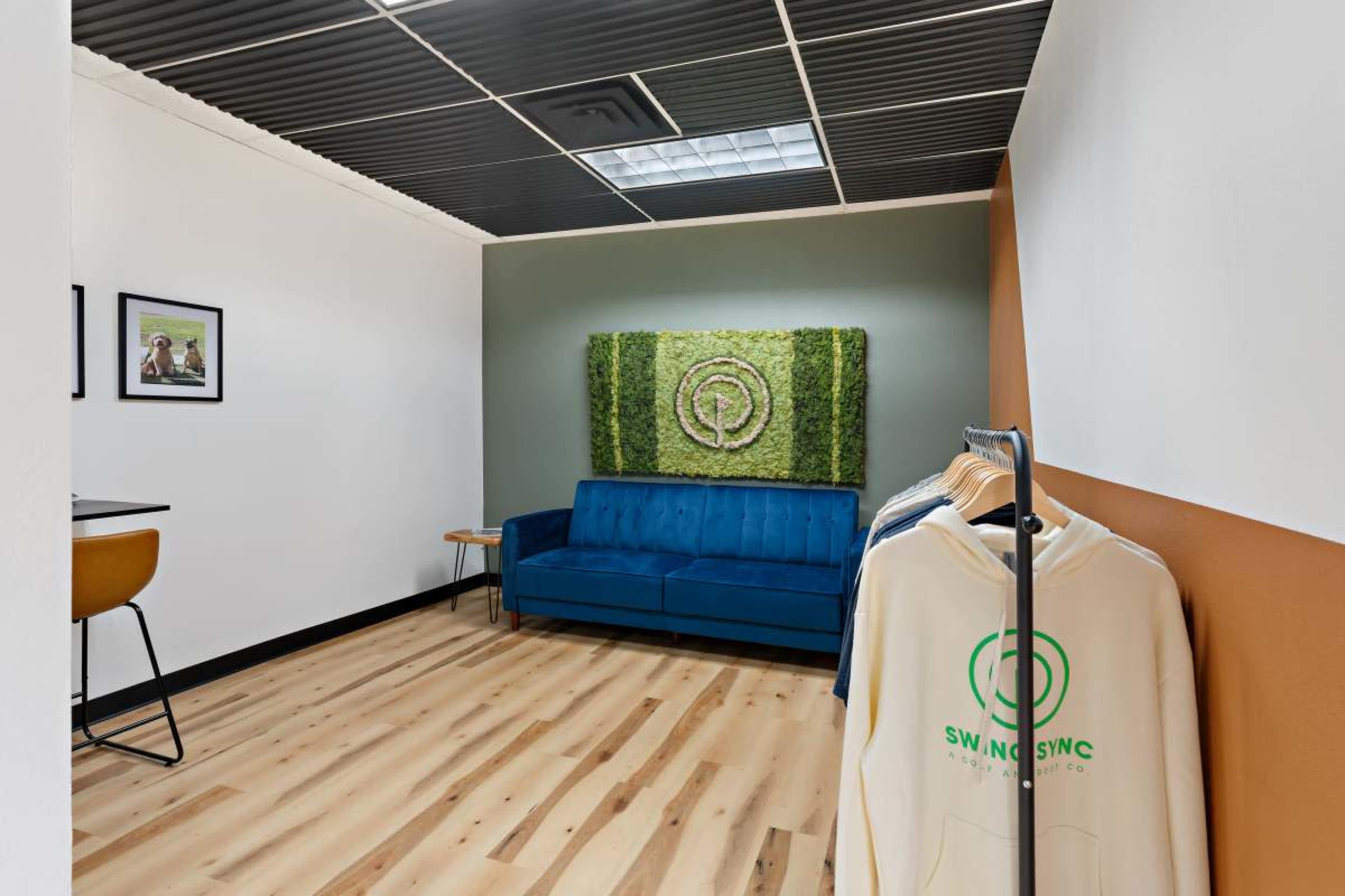 The image shows a modern office interior featuring a blue couch, a green wall art piece, and a clothing rack with hoodies.