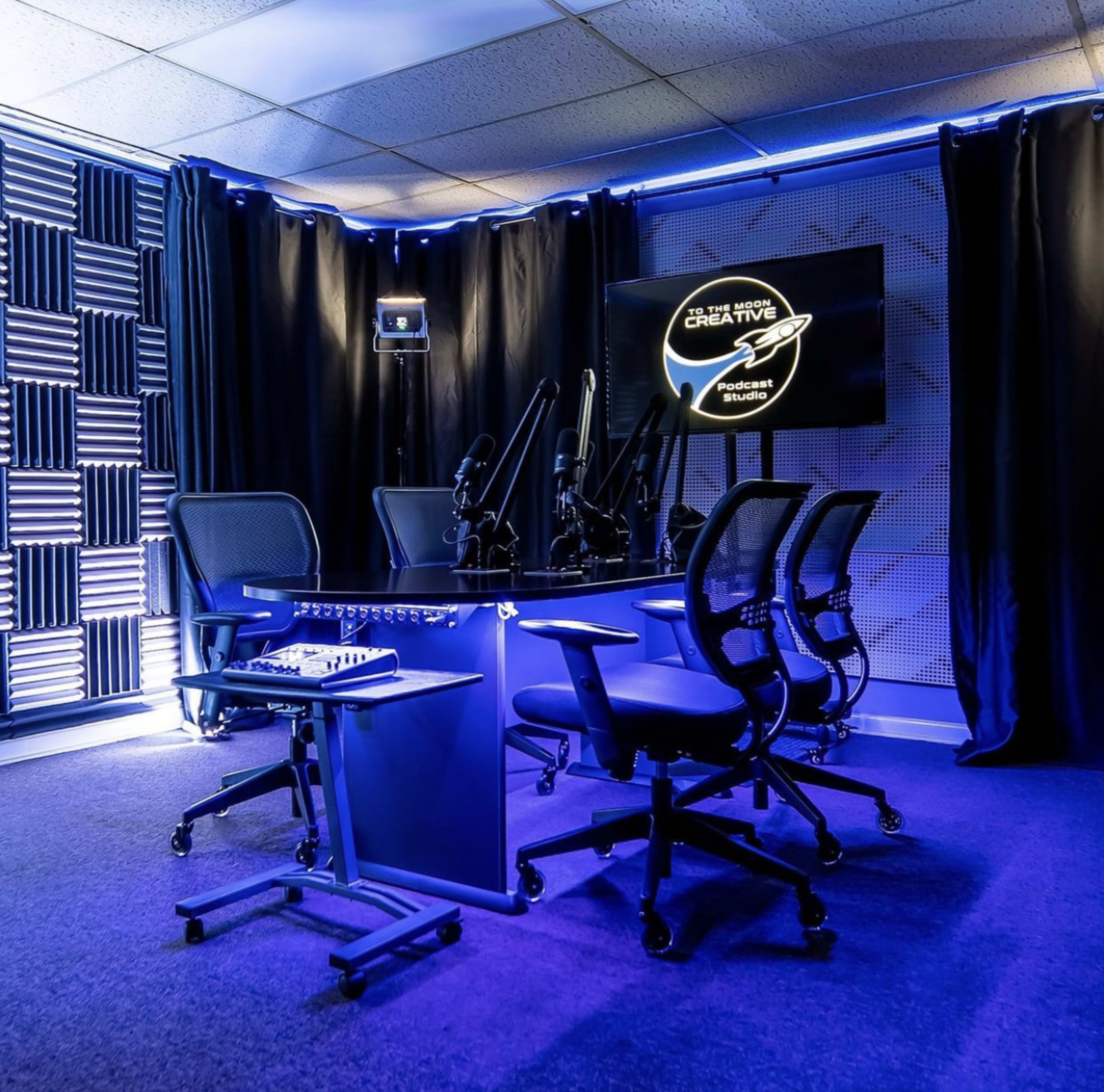 A modern podcast studio featuring a round table with microphones, chairs, and acoustic paneling, illuminated by blue lighting.