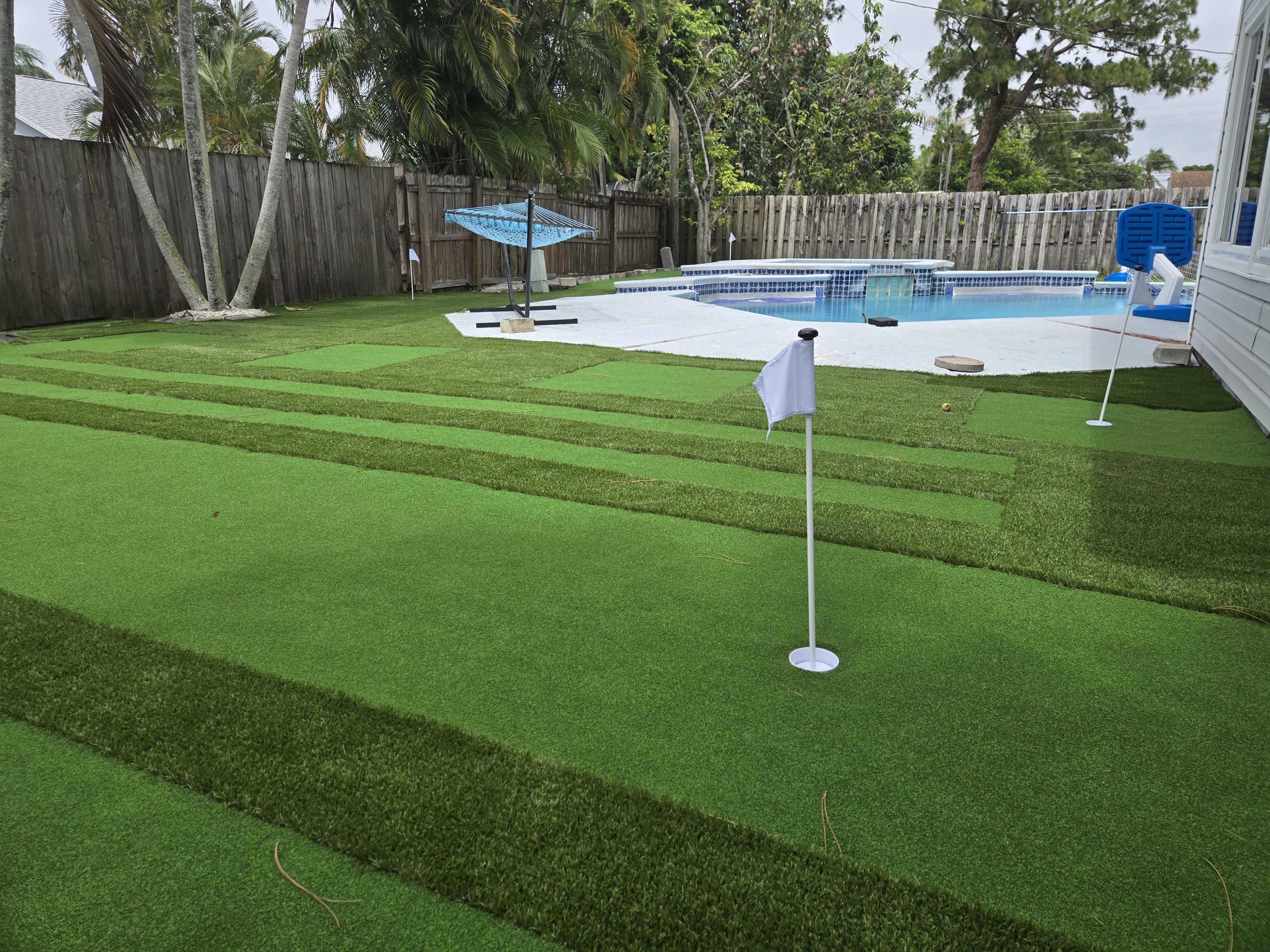 The image shows a backyard with artificial grass featuring distinct green stripes and a small putting green, alongside a swimming pool and palm trees in the background.