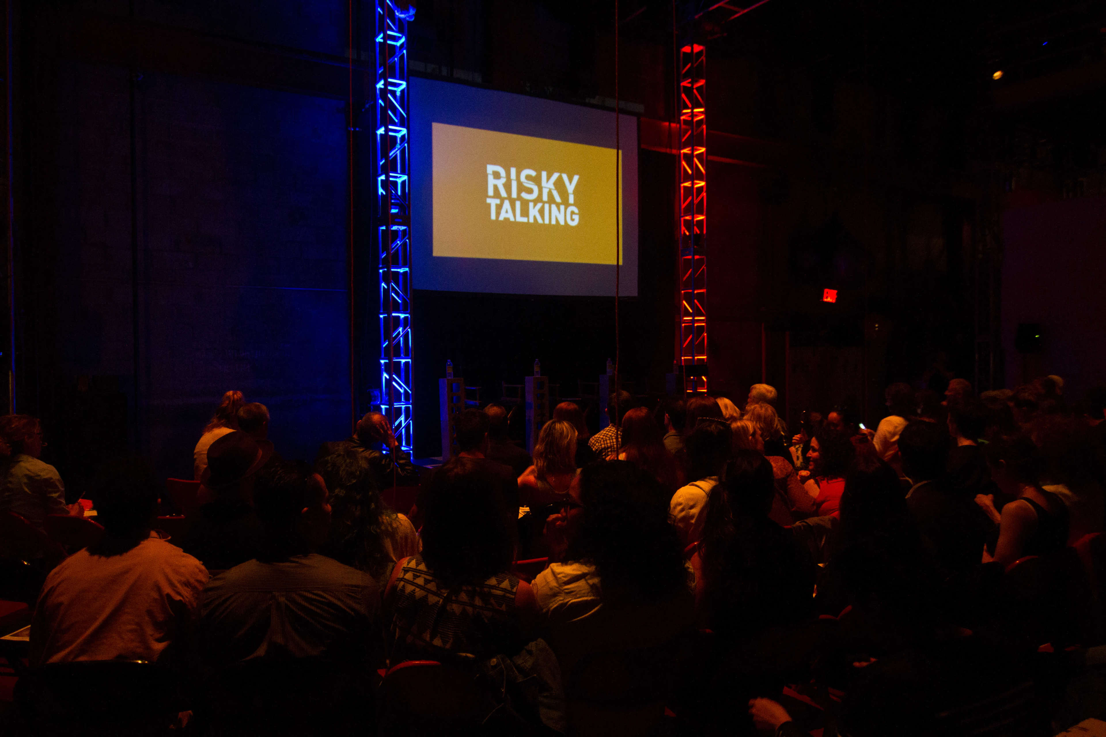 A dimly lit auditorium filled with an audience seated in rows, facing a large screen displaying the words "RISKY TALKING."