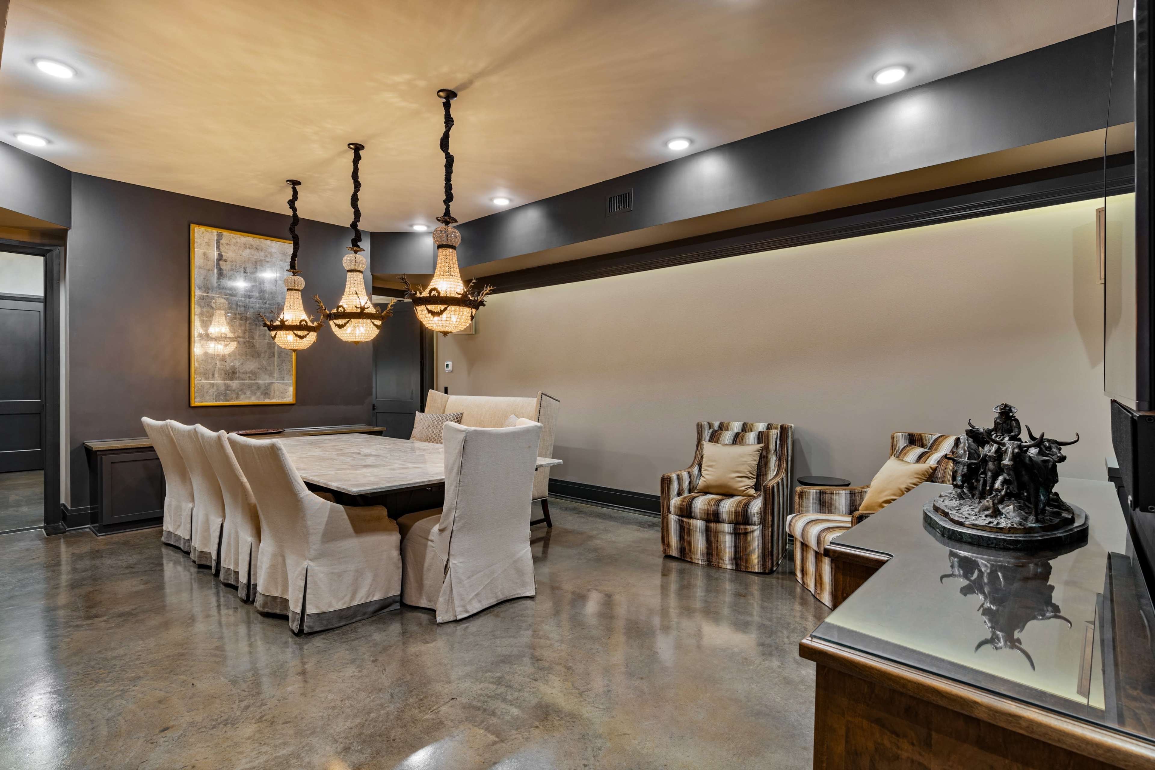 The image shows a modern dining room featuring a large table surrounded by white upholstered chairs, with a statement light fixture overhead and a decorative sculpture on a nearby cabinet.