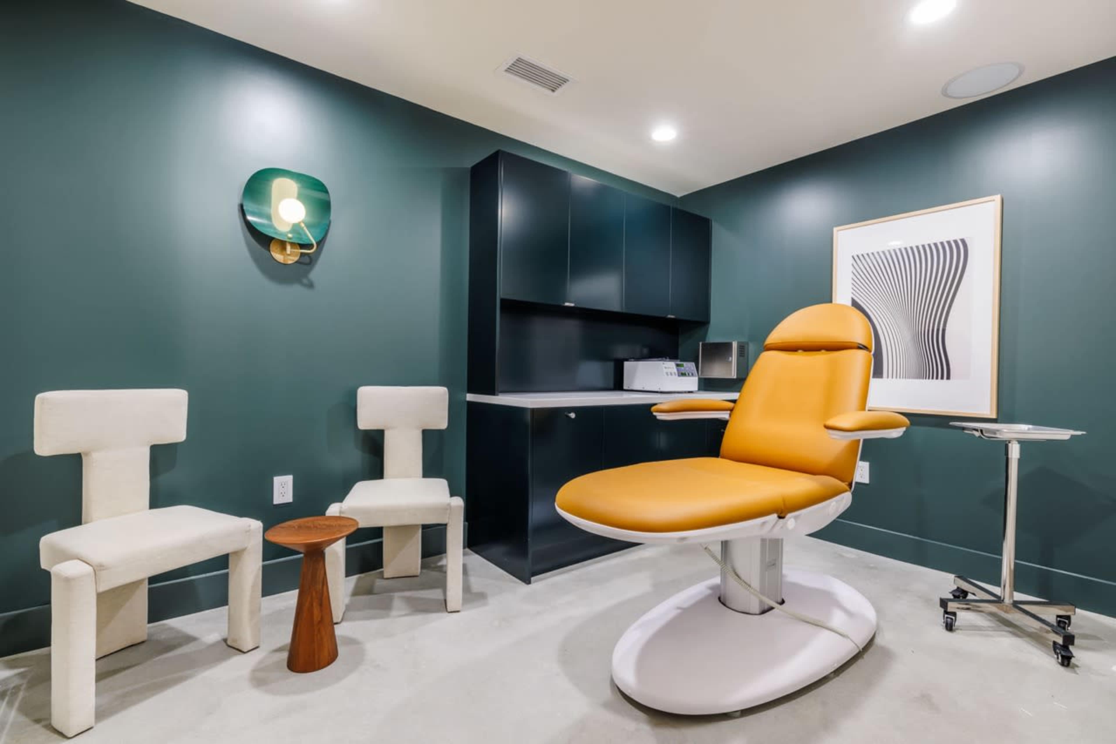 A medical examination room features a bright yellow chair, two white armchairs, and a dark green wall with a wall-mounted light fixture.