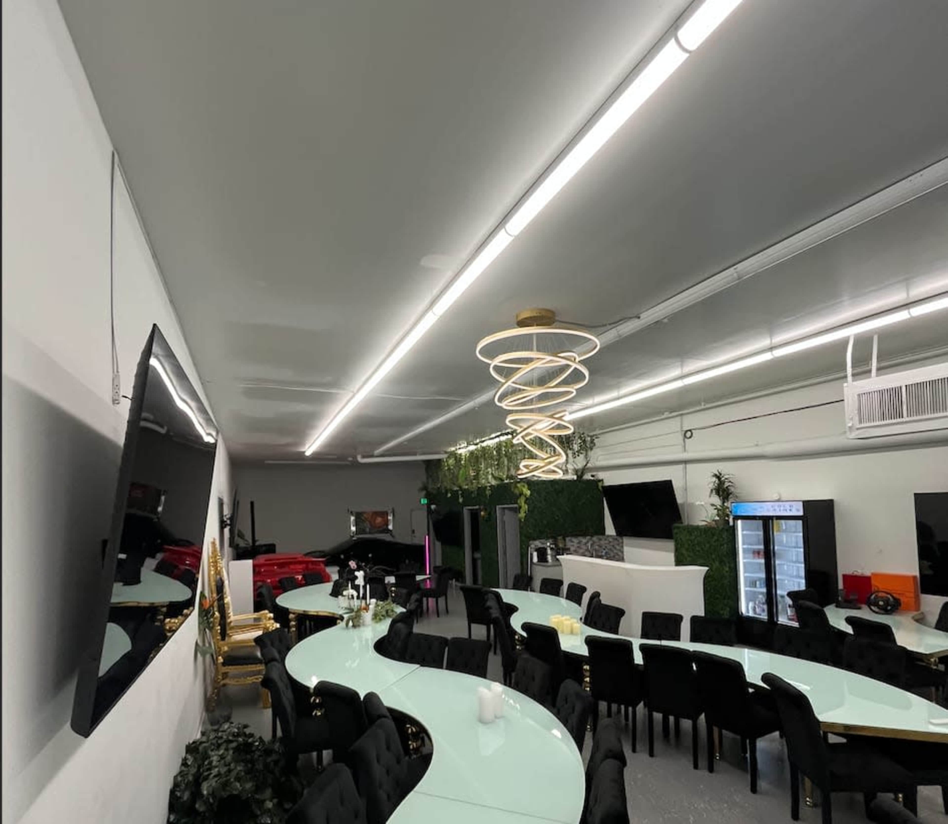 The image shows a modern dining area with a long, curved table, black chairs, and a spiral light fixture, alongside walls adorned with large televisions and greenery.