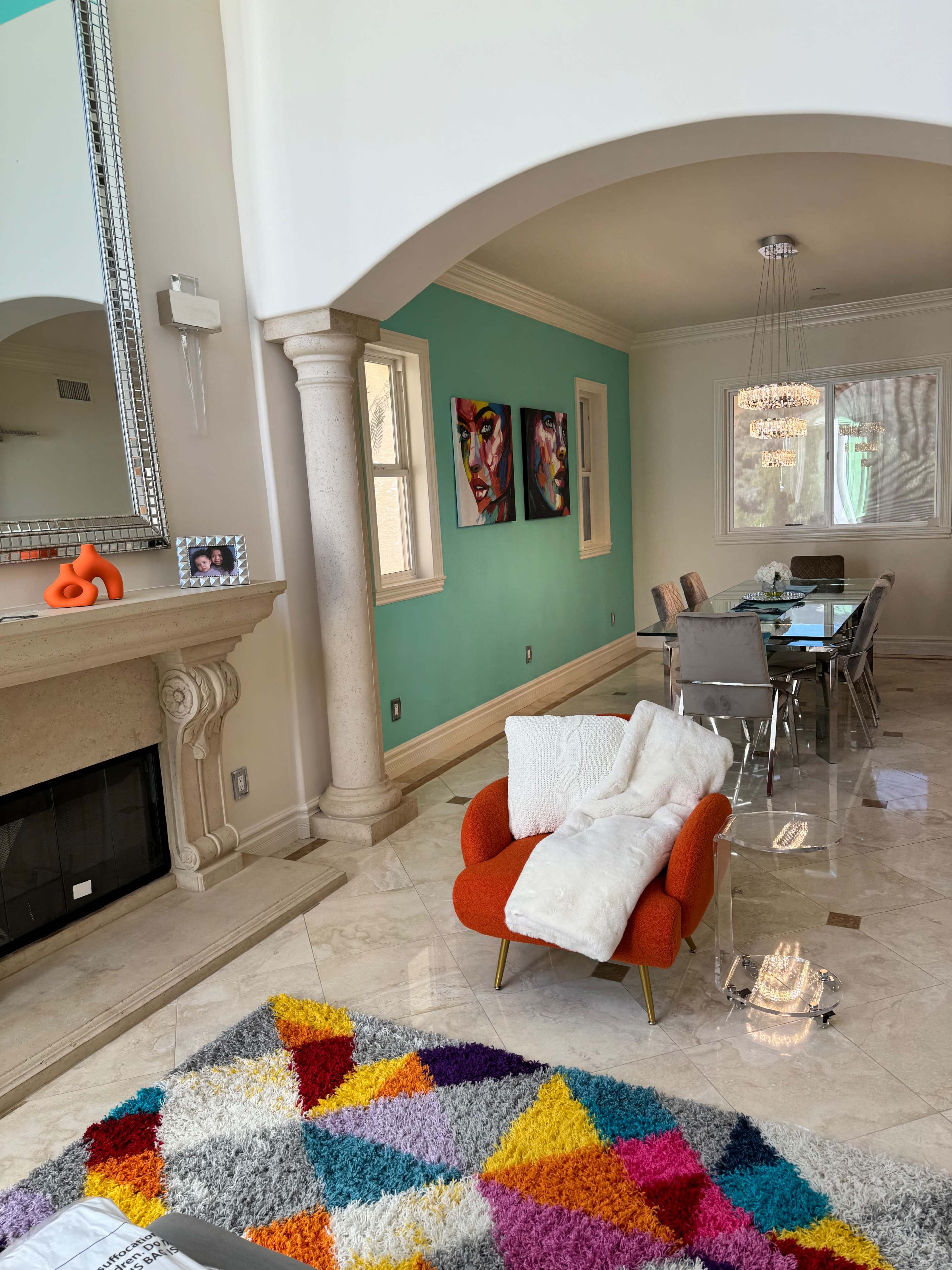 The image shows a modern living space featuring a bright orange chair, a colorful patterned rug, a glass dining table, and artwork on the turquoise wall.