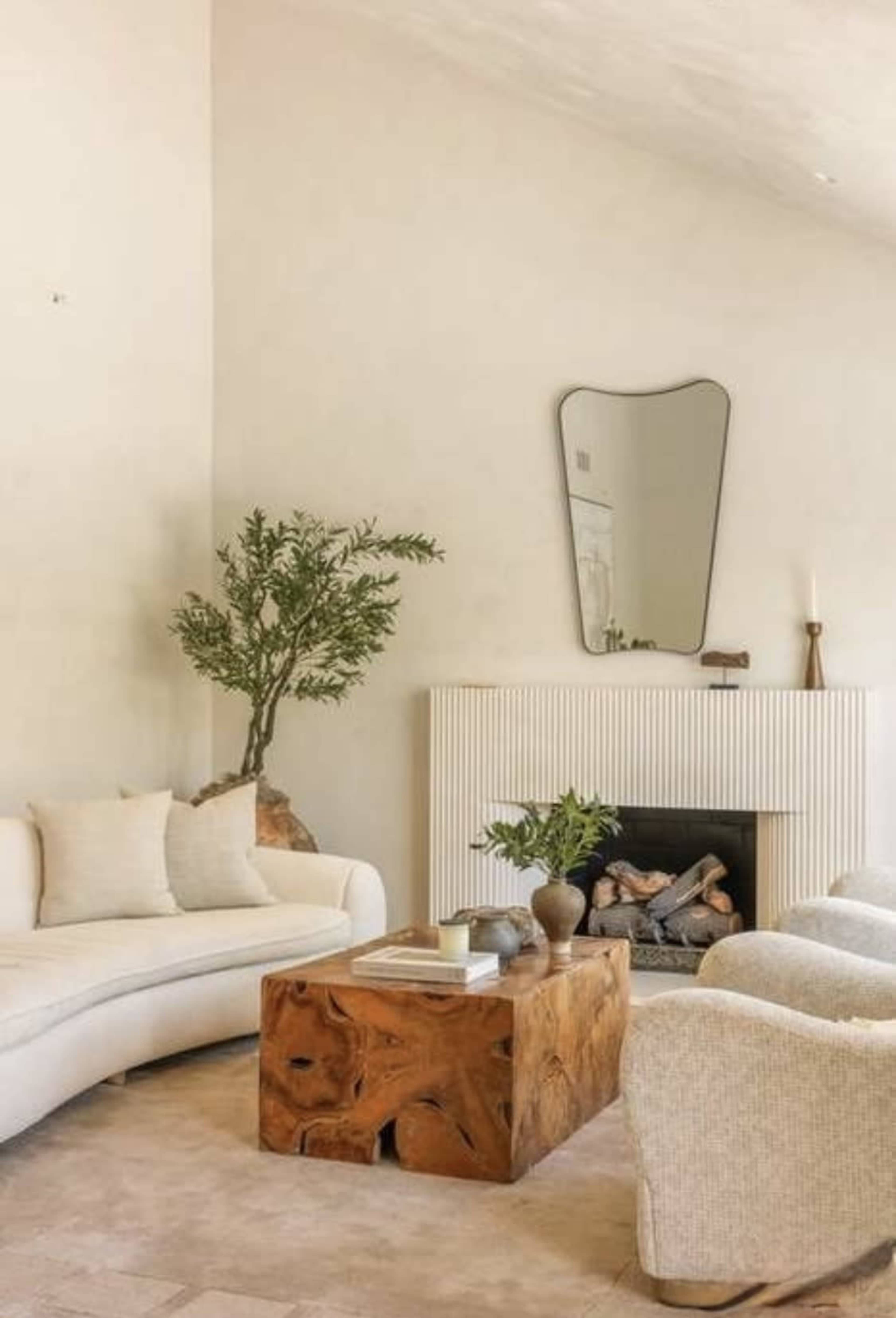 The image depicts a minimalist living room featuring a curved white sofa, a wooden coffee table, and two textured armchairs, with a fireplace and a large mirror on the wall.