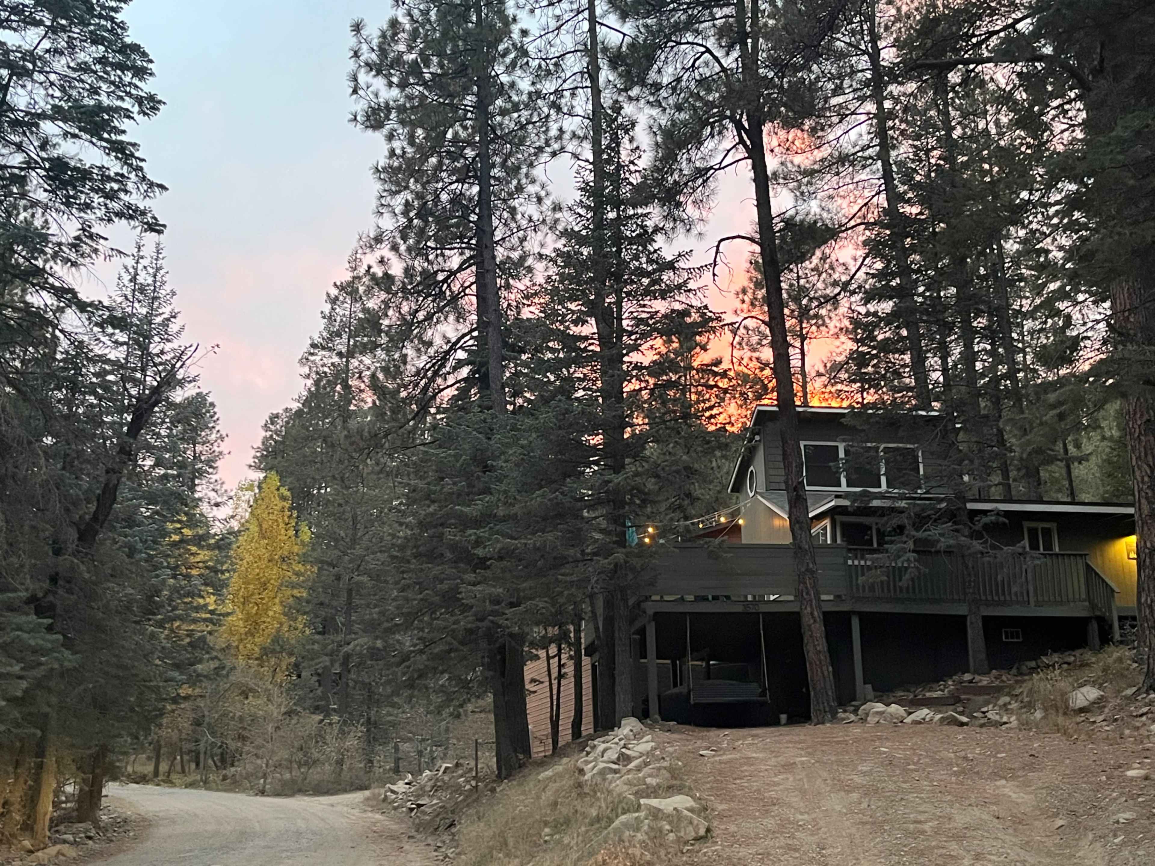 A two-story house is nestled among tall pine trees along a gravel road, with a sunset glow visible in the background.
