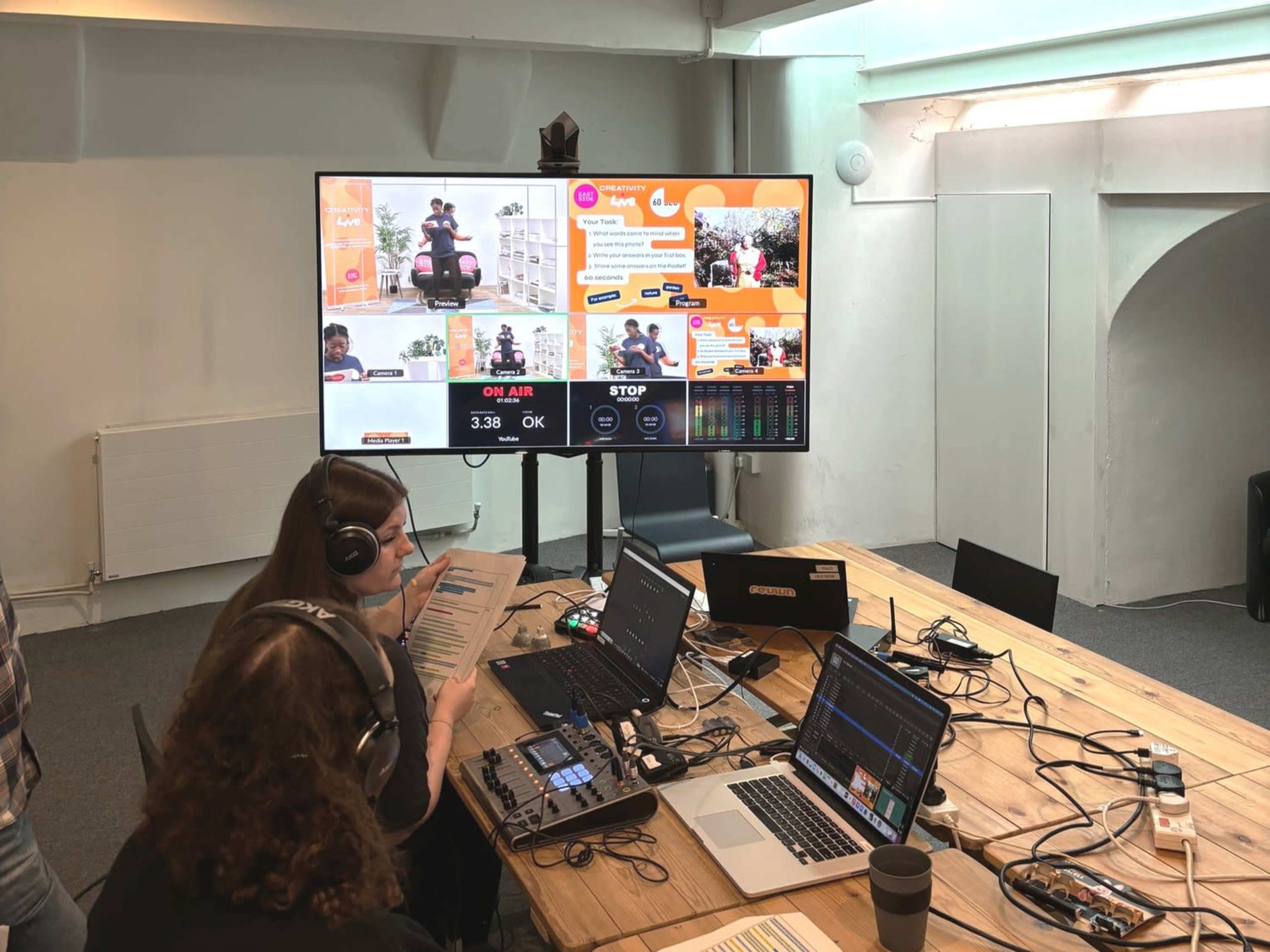 A person sits at a table with multiple laptops and audio equipment, monitoring a digital broadcast on a large screen in a bright, modern workspace.