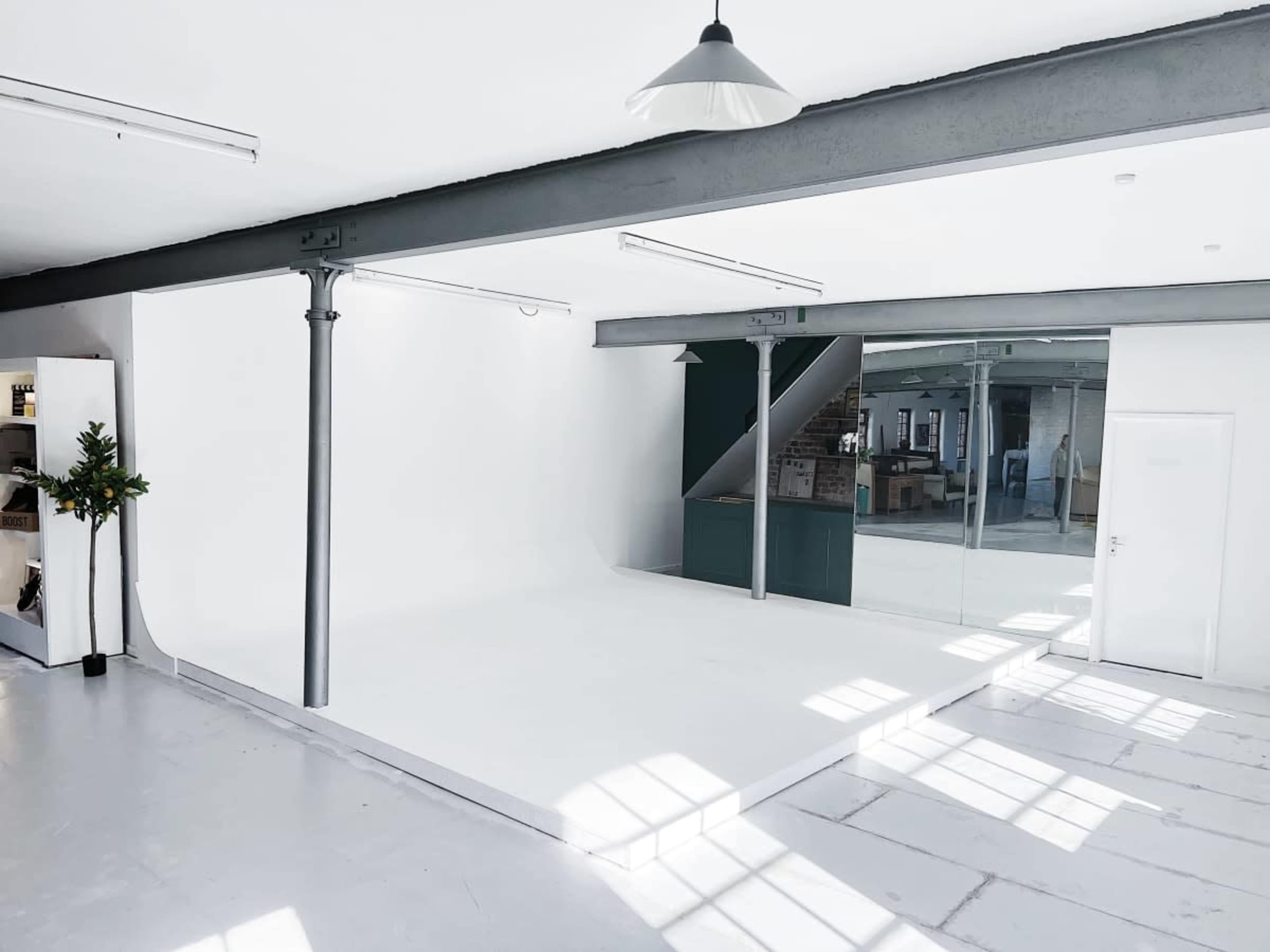 A bright, minimalist studio space with white walls and floor, featuring large windows and industrial-style columns.
