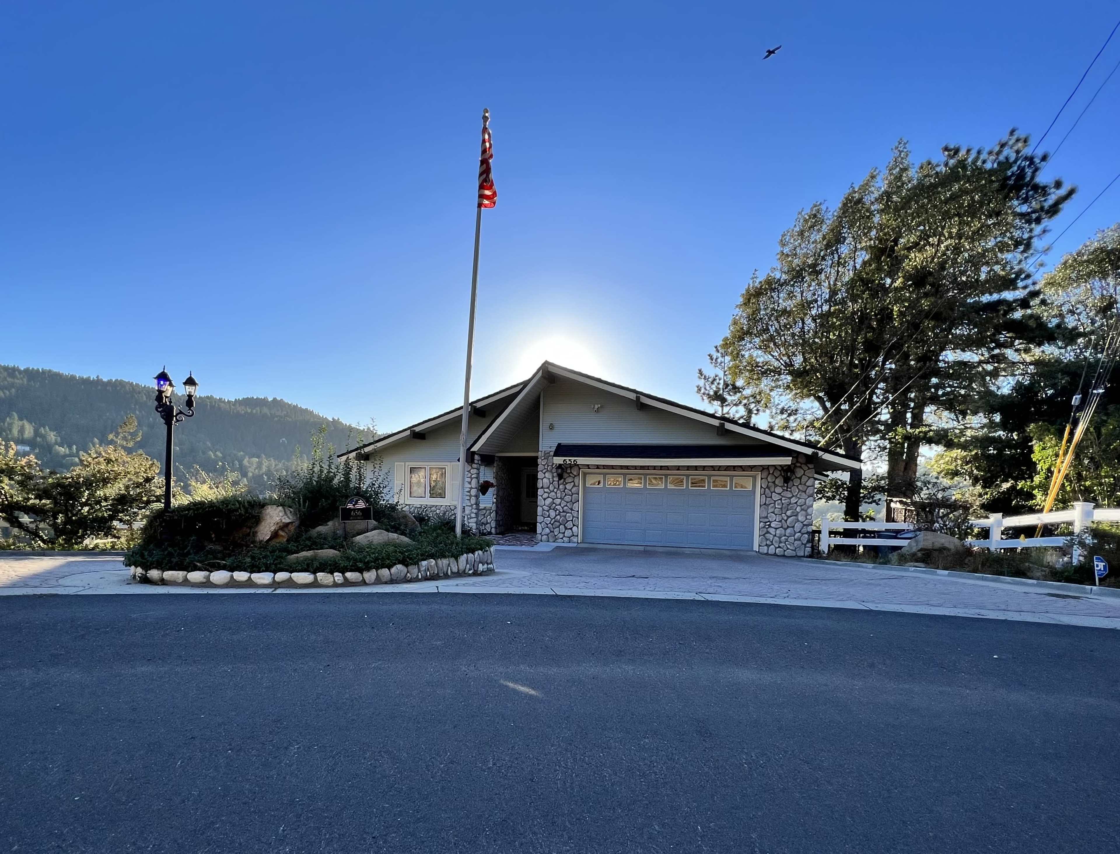 A single-story house with a stone facade and an attached garage is situated at the end of a curved driveway, surrounded by trees and a mountain backdrop.