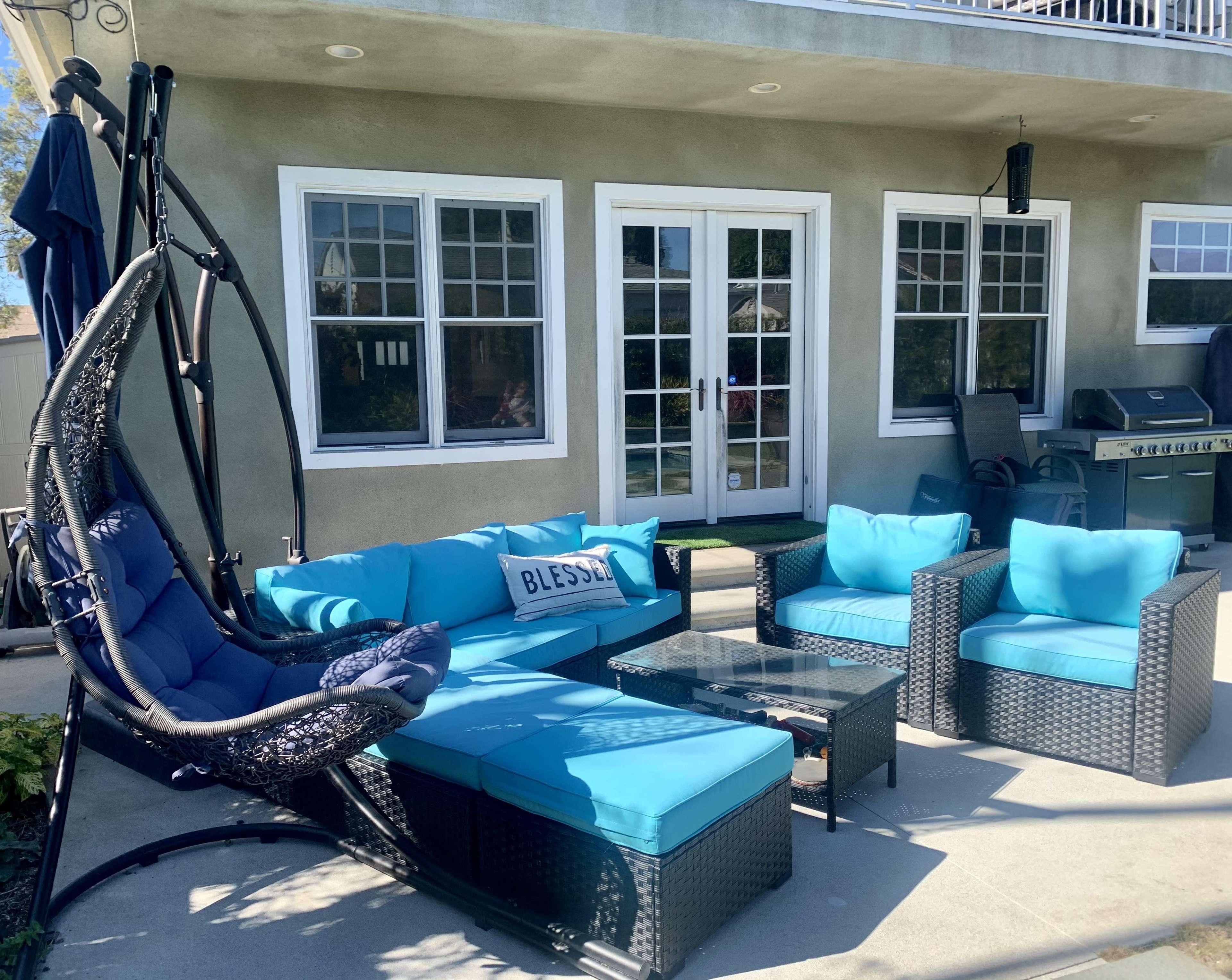 A patio area features a turquoise seating arrangement, including a hanging chair, a sectional sofa, and a coffee table, in front of a set of large windows and doors.
