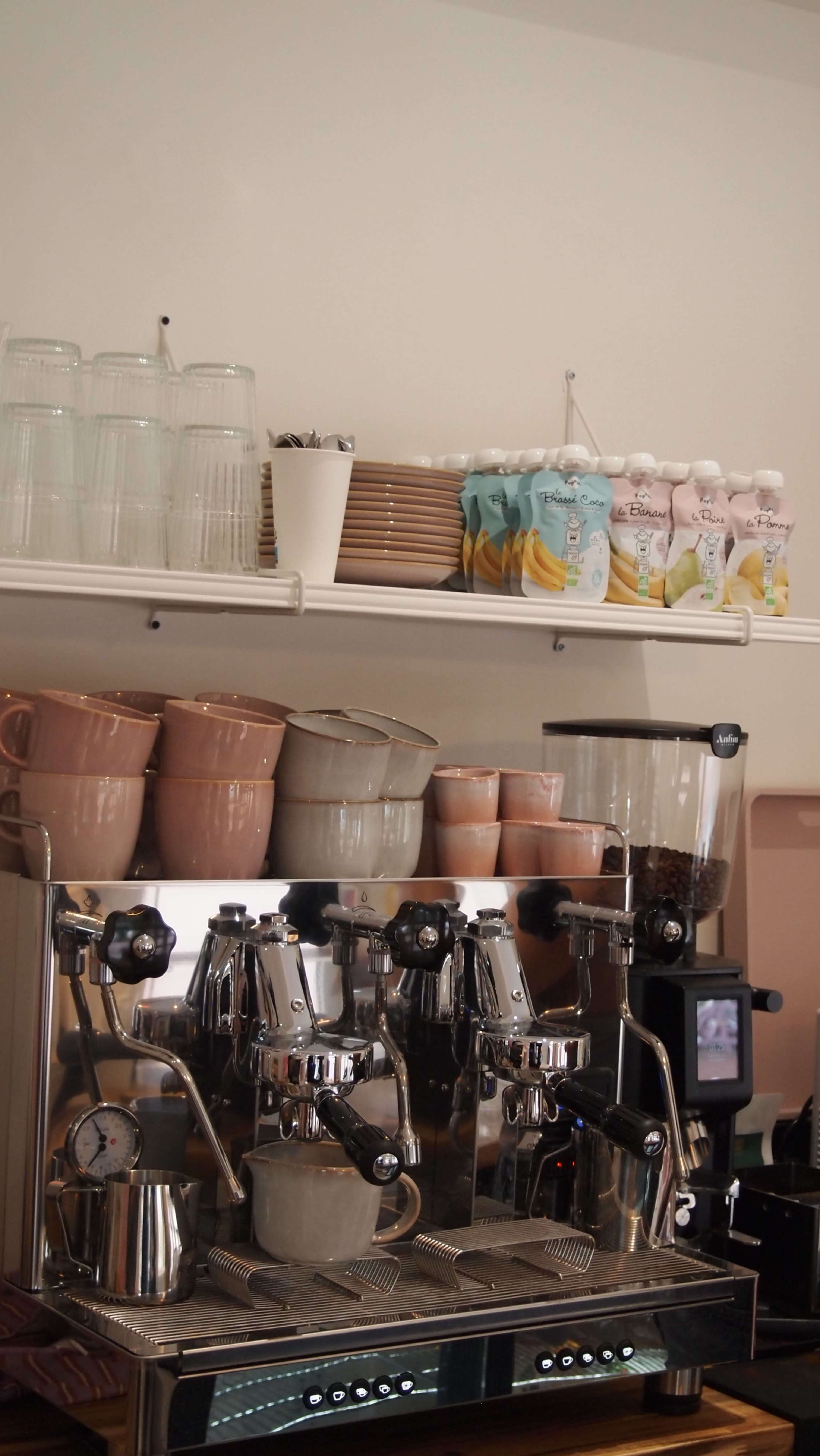 A coffee machine is displayed on a countertop with various ceramic cups, glassware, and milk cartons arranged on shelves above.