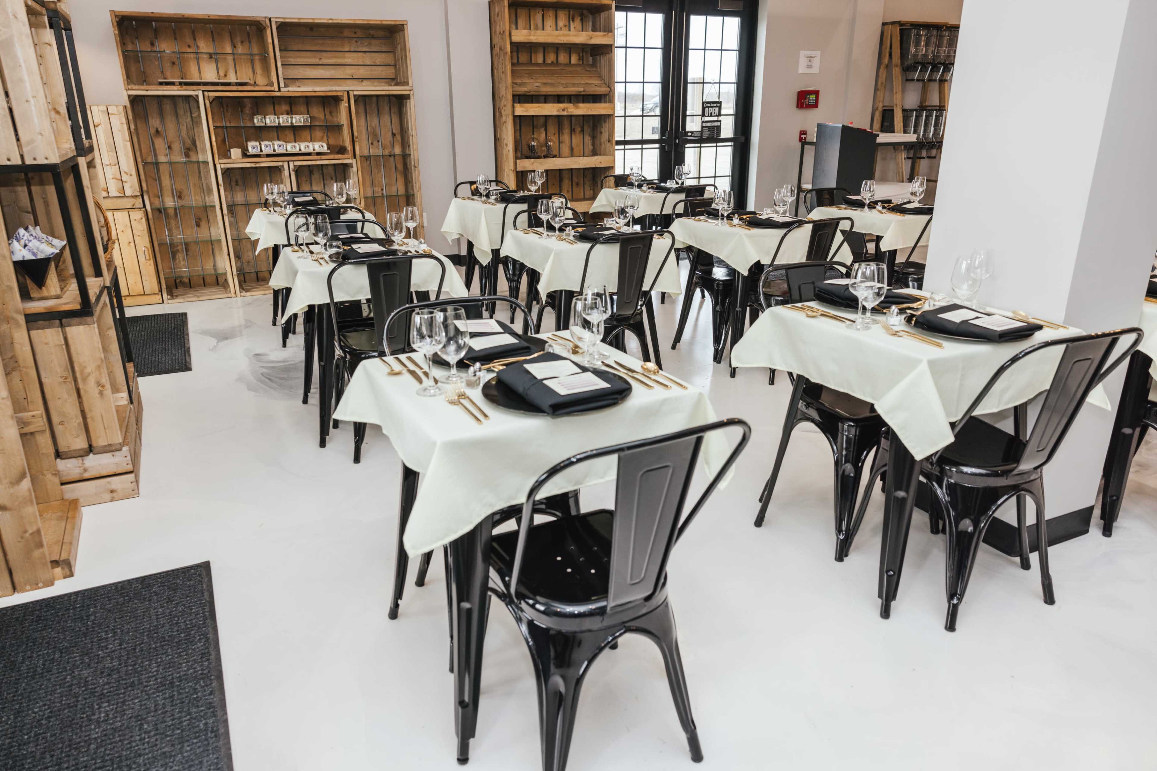 The image shows a dining area with several tables set with white tablecloths, black chairs, and neatly arranged utensils in a modern, wooden interior.