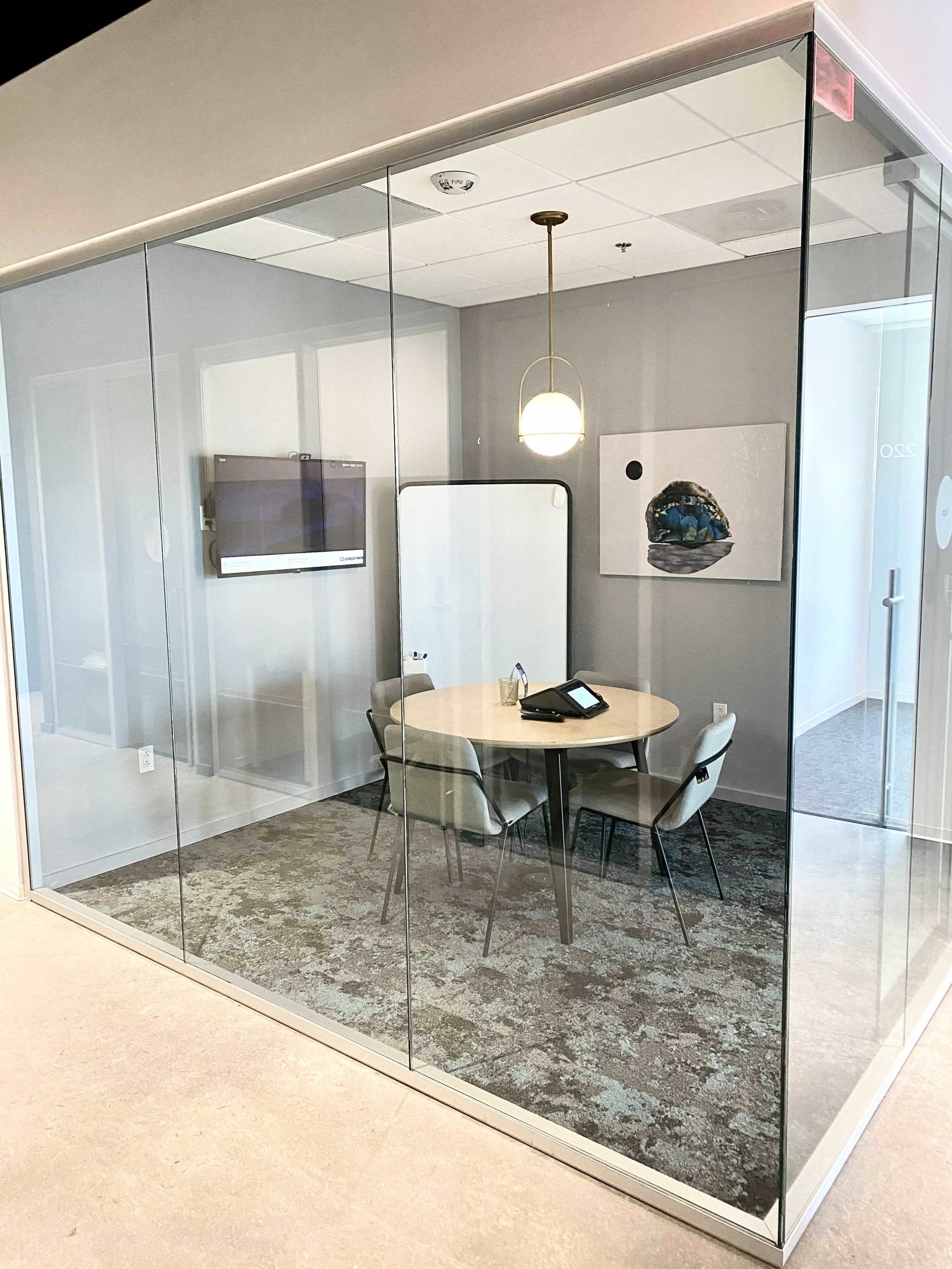 A modern conference room enclosed by glass walls, featuring a round table surrounded by chairs and a presentation screen on one wall.