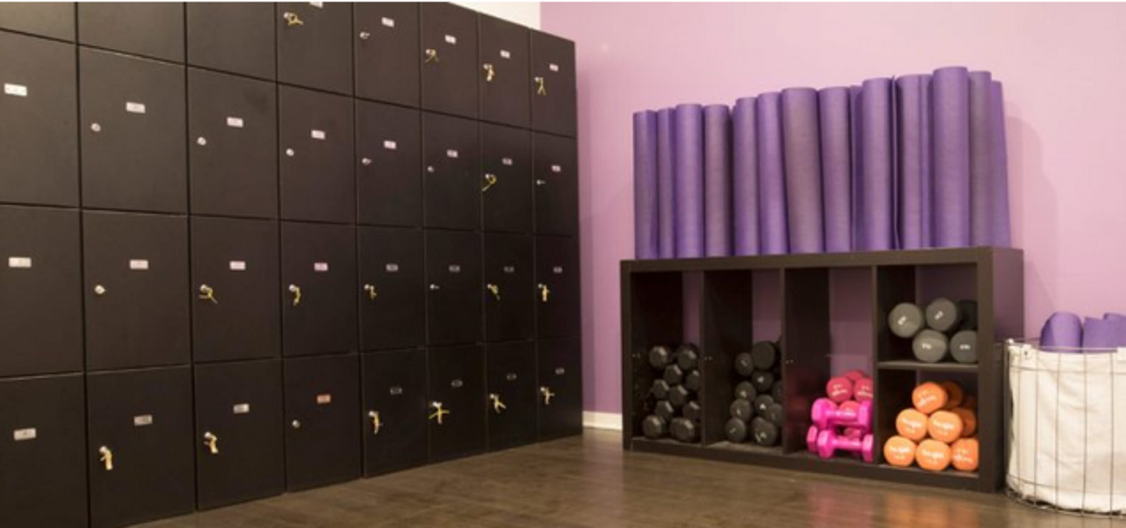 The image shows a fitness room with black lockers on one wall and neatly arranged purple yoga mats and weights on shelves.