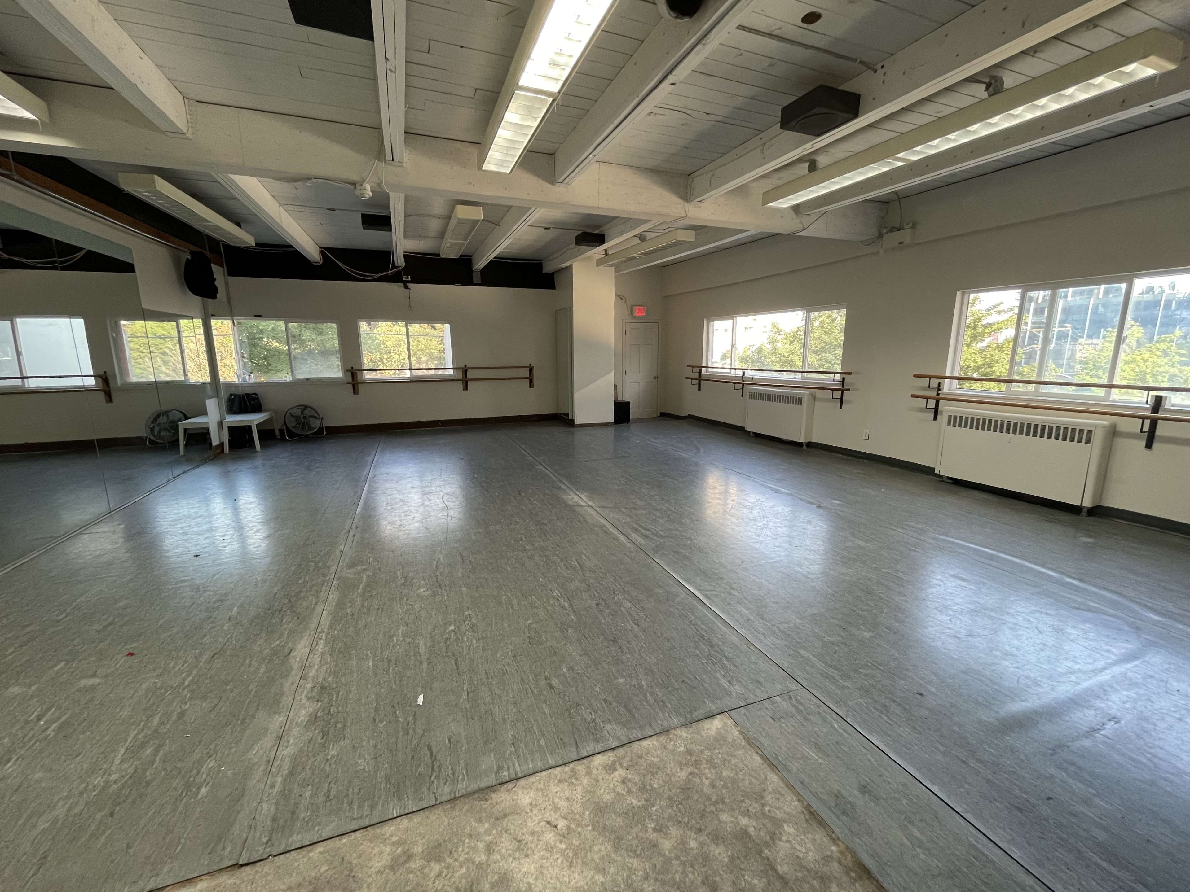 The image shows a spacious dance studio with wooden ballet barres along the walls, large windows allowing natural light, and gray flooring.