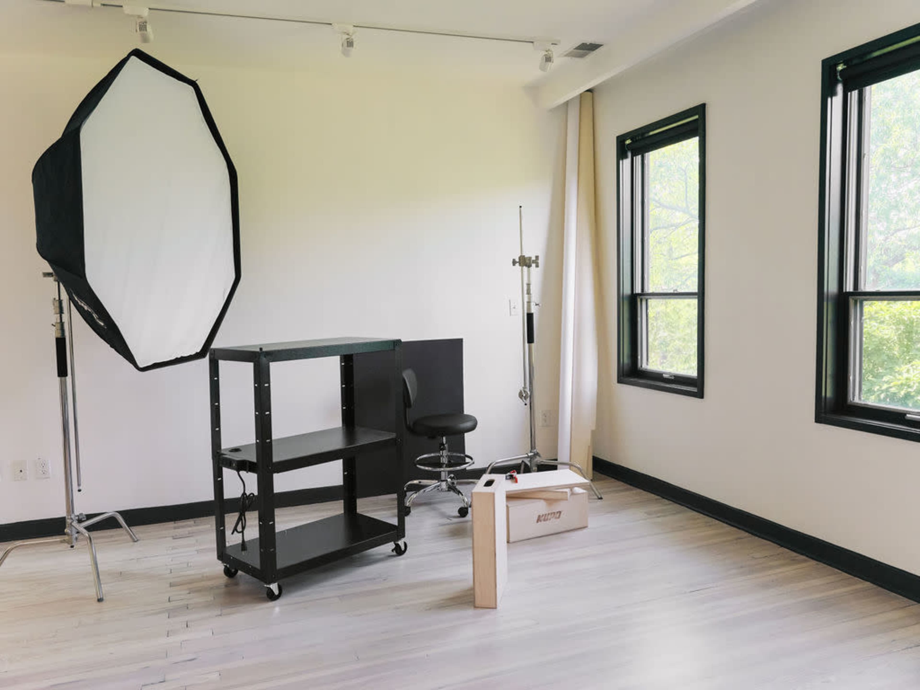 The image depicts a bright, empty studio space featuring a large softbox light, a black rolling cart with shelves, a swivel chair, and a wooden crate on the floor.
