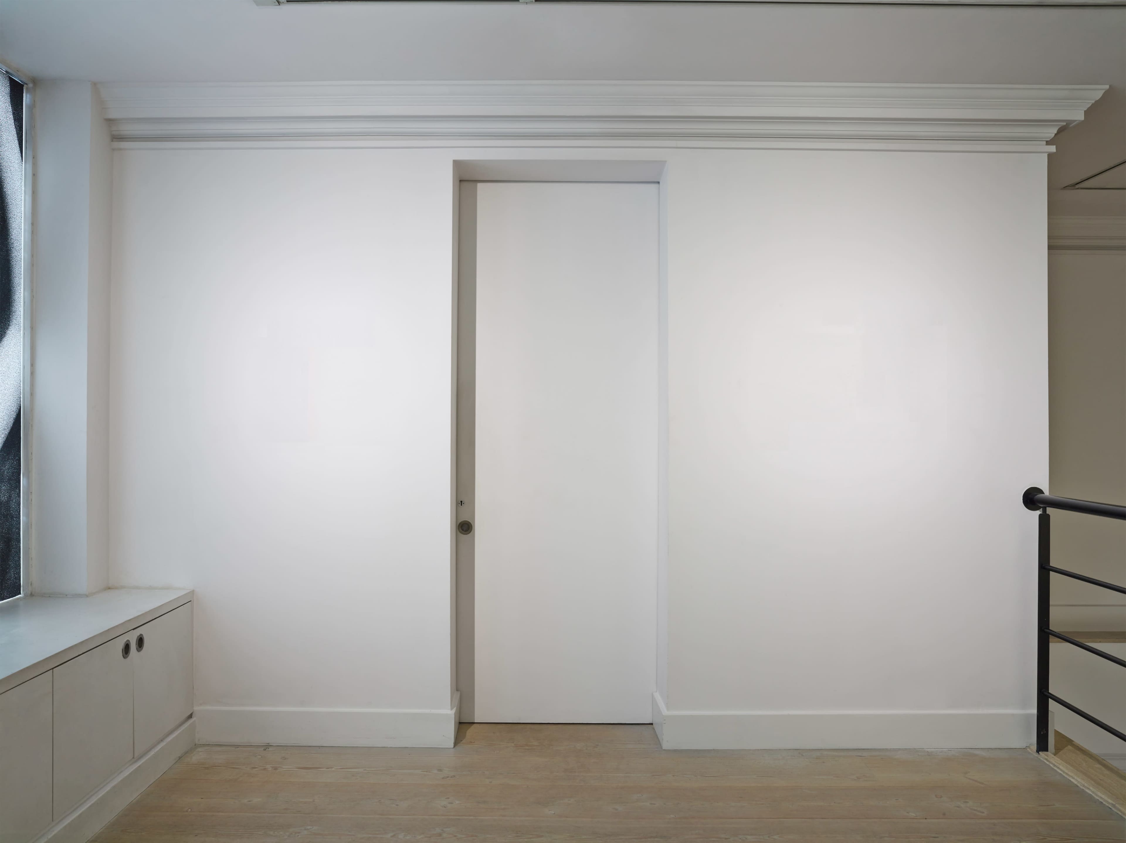 A plain white wall features a concealed door and a window with a view.