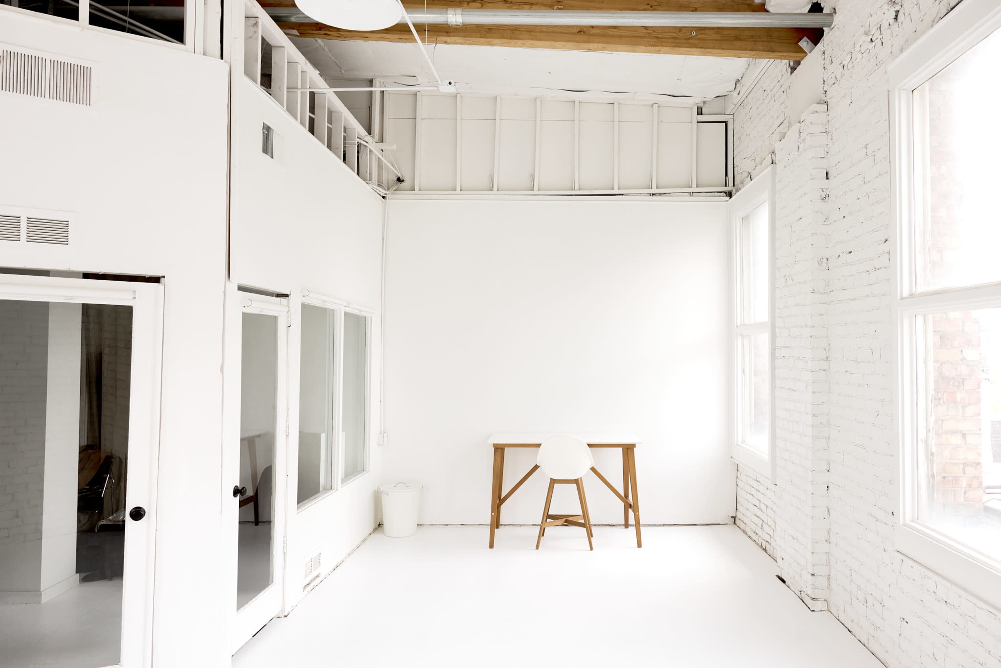 A minimalistic white room with a wooden table and a single chair, featuring large windows and exposed brick walls.