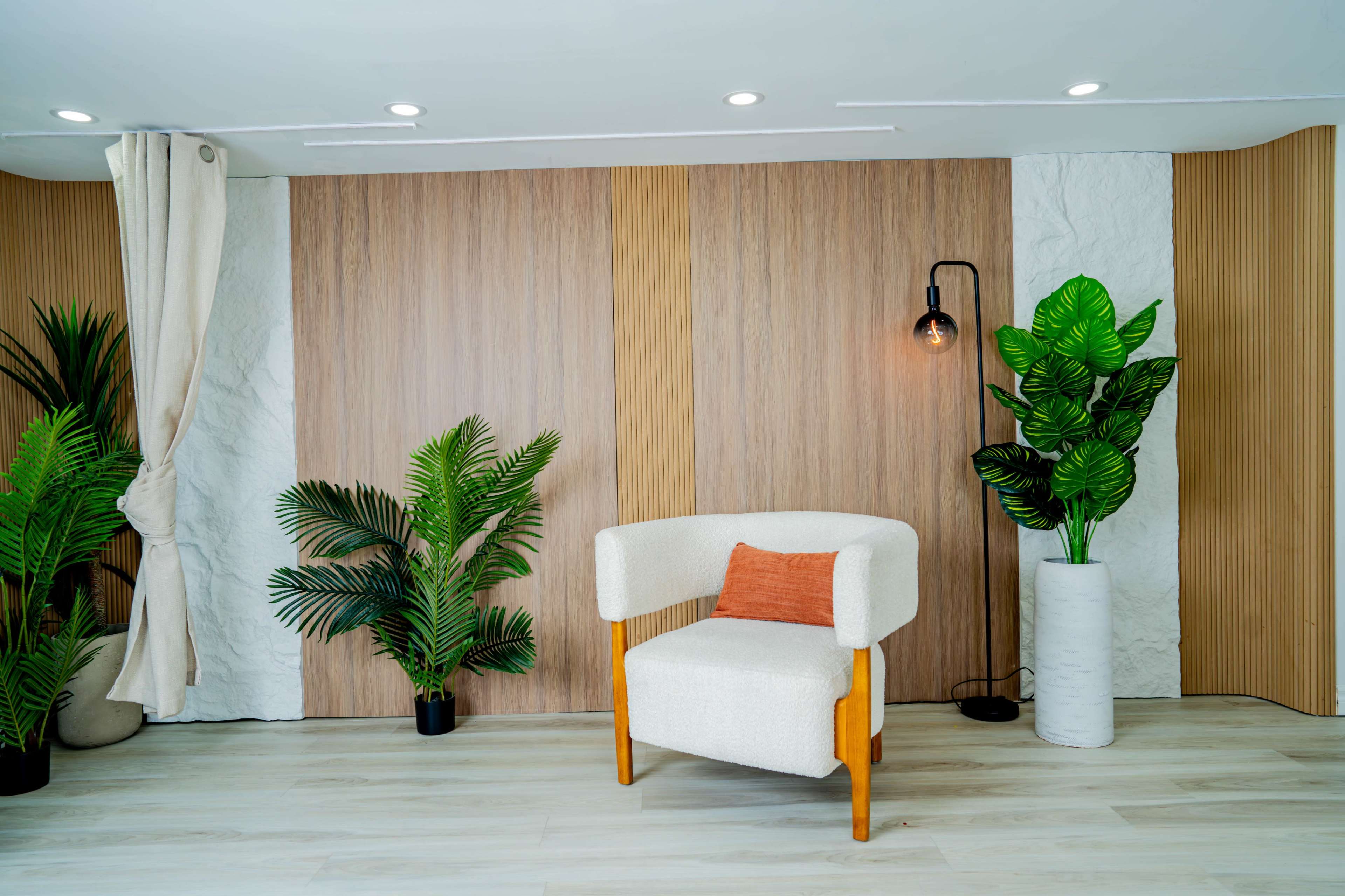 The image shows a modern living space featuring a single upholstered chair with a decorative pillow, surrounded by potted plants and a wooden accent wall.